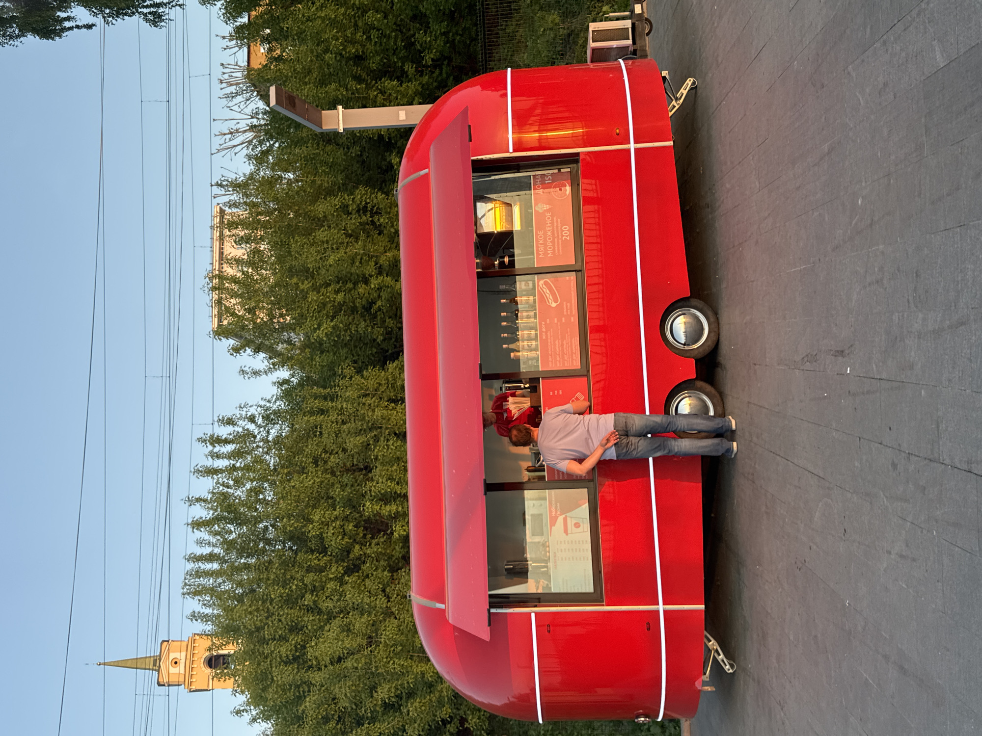 The image depicts a bright red, modified food truck resembling a classic British double-decker bus, located outdoors. A man is standing at the service window of the truck, possibly ordering food. The food truck features white decorative stripes and windows displaying menu items and prices in Russian. The surrounding environment includes greenery, a church with a spire, and overhead power lines. The image was likely taken in the city of Perm, Russia.