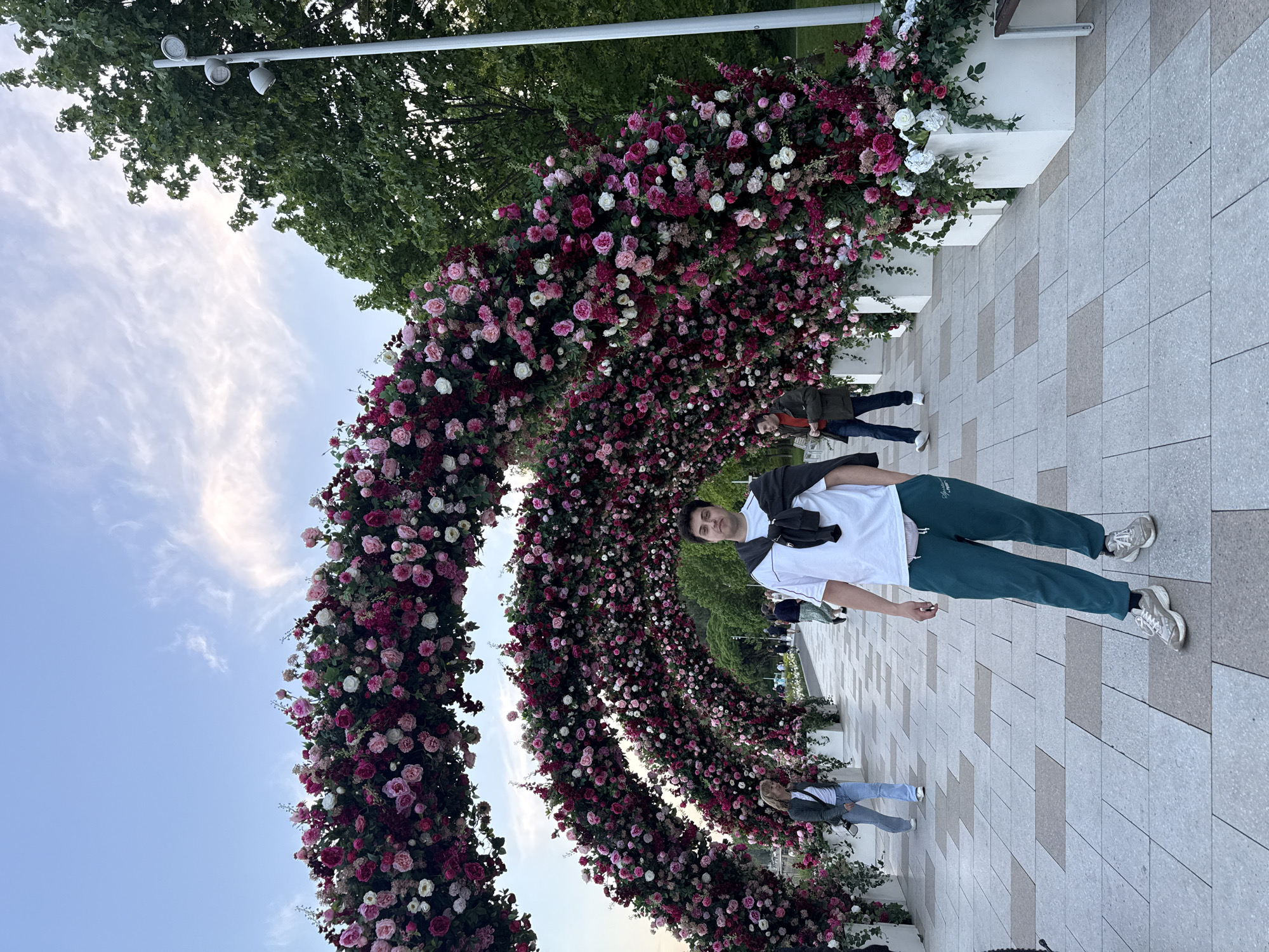 A man stands in front of a large flower archway on a tiled walkway. The archway consists of pink, white, and deep red roses and foliage. He is wearing a white T-shirt, green sweatpants, and gray sneakers, and has a black jacket draped over his left shoulder. The sky is blue with some clouds. Trees and lampposts are visible in the background. The setting appears to be a public space in Moscow, Russia.