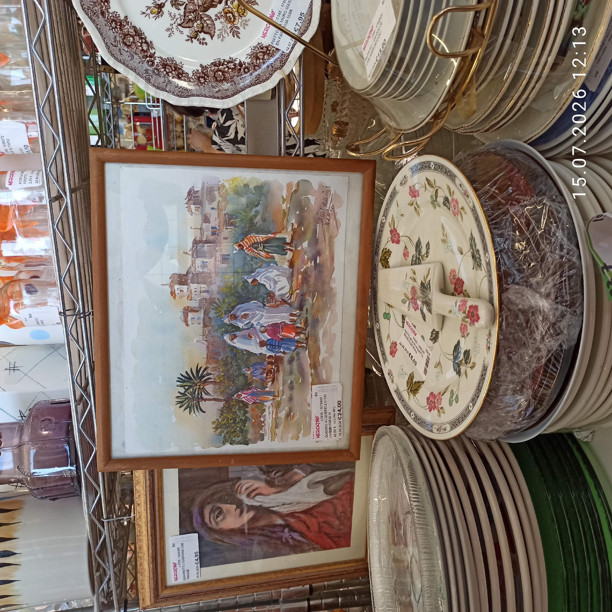The image shows a retail shelf display at a mercatino (second-hand market) in Molfetta, Italy. The shelves are filled with various items, primarily dishes and decorative objects. In the foreground, a stack of white plates with a floral pattern is visible, topped with a cake server. To the left, a framed watercolor painting of a woman with a purple headscarf is displayed, with a price tag indicating "€4,95". To the right of that, another framed watercolor depicts a scene with figures in robes and palm trees, also with a price tag of "€24,00". Behind these paintings, various glass jars and vases are arranged on higher shelves. In the background, more stacks of plates are visible, some with decorative rims. The overall scene suggests a bustling marketplace environment. The date stamp "15.07.2026 12:13" is superimposed on the bottom right corner of the image.