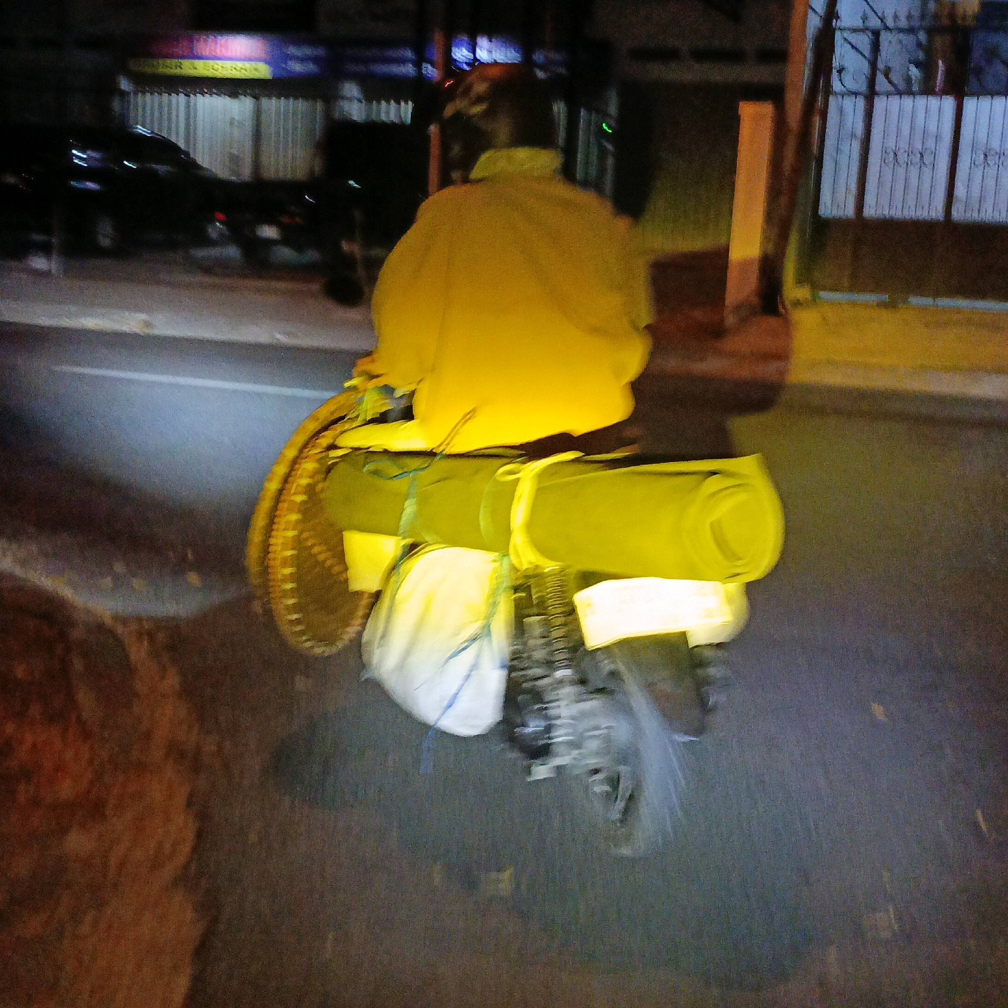 This is a nighttime, motion-blurred photograph of a person riding a motorcycle laden with cargo in Salatiga, Indonesia. The rider is wearing a bright yellow jacket and a helmet. Attached to the rear of the motorcycle are two large, rolled items secured with straps, possibly sleeping bags or camping mats, and a white bundle, which could be additional luggage. The motorcycle also appears to have a woven basket attached to the side. The background shows a street scene at night, with the blurred lights of buildings and vehicles. On the right side of the image, a metal gate with vertical bars and a sign above it are visible. The street surface is dark asphalt, with some reflections from streetlights. The image conveys a sense of movement and a journey in progress.