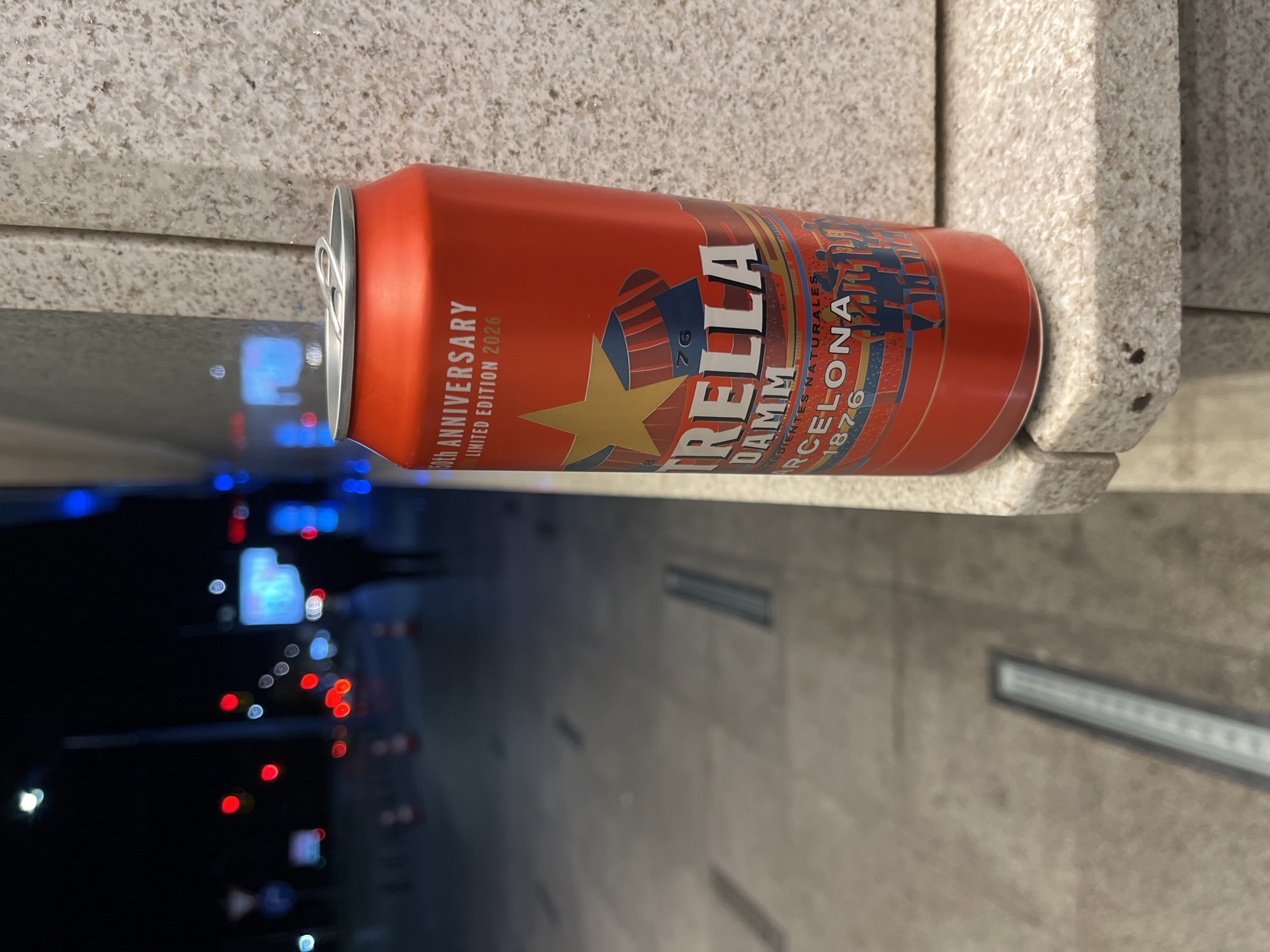 A 60th Anniversary Limited Edition 2026 can of Estrella Damm Barcelona 1876 beer rests horizontally on a light-colored ledge of an urban structure. The background, visible through the reflective surface under the can, appears to be a street at night, illuminated by streetlights and vehicle lights. The ledge shows signs of wear and weathering. The location is presumed to be Sofia, Bulgaria, based on the prompt.