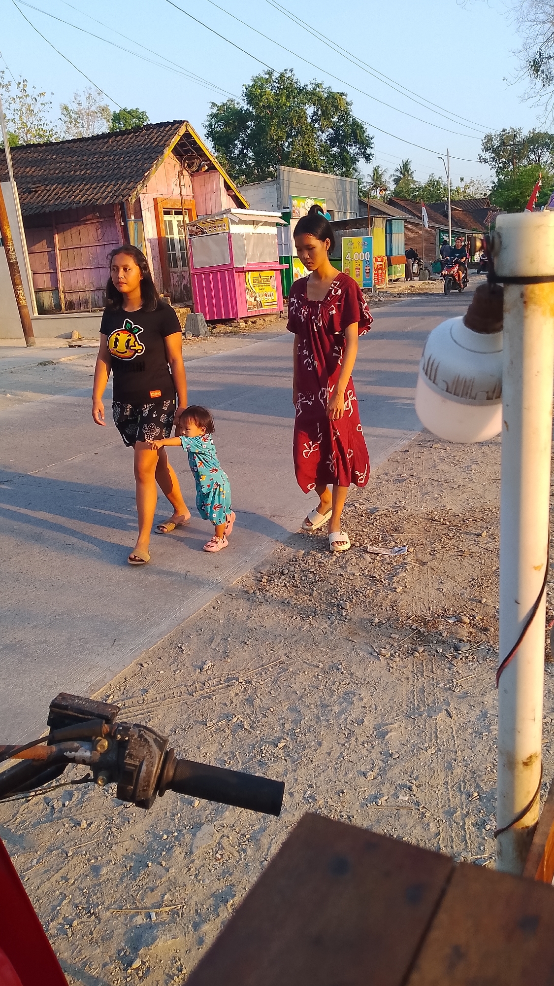 In the city of Karanglangu, Indonesia, two women and a child are walking along a dusty road. The woman on the left wears a black t-shirt with an orange design and black shorts. The woman on the right is wearing a long, red, patterned dress. The child, dressed in a blue, patterned outfit, walks between them, reaching out to touch the leg of the woman in black. In the background, small buildings with colorful facades line the road. The sun is low in the sky, casting long shadows. A motorcycle is visible in the distance, and part of a vehicle's handlebars is in the foreground, suggesting the photo was taken from a stationary position.