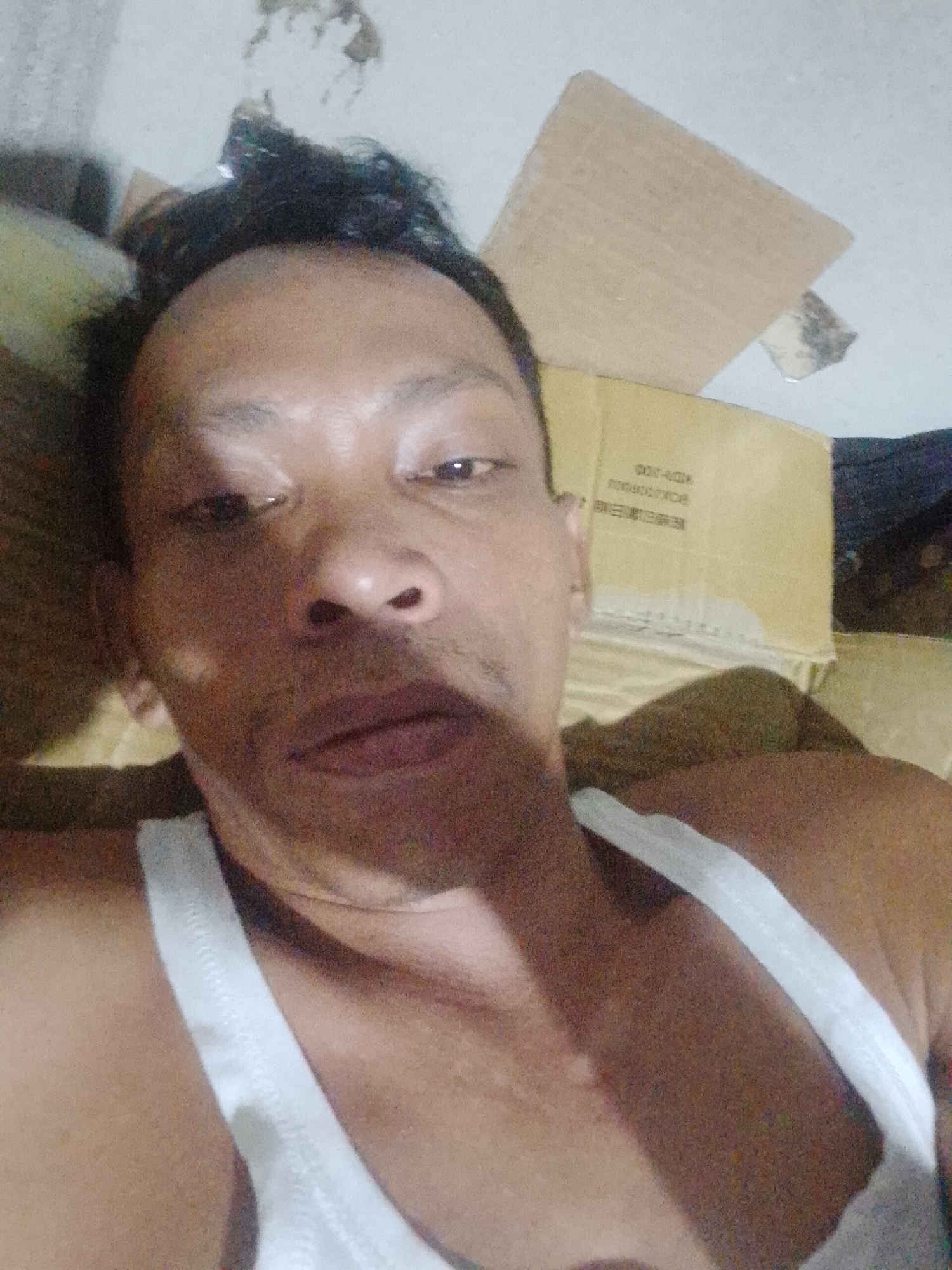 A close-up photograph of a middle-aged Asian man wearing a white tank top is presented. The man is lying down, his head positioned at the top of the frame. Behind him are two cardboard boxes. The photograph was taken in Bukit Ketok, Indonesia.