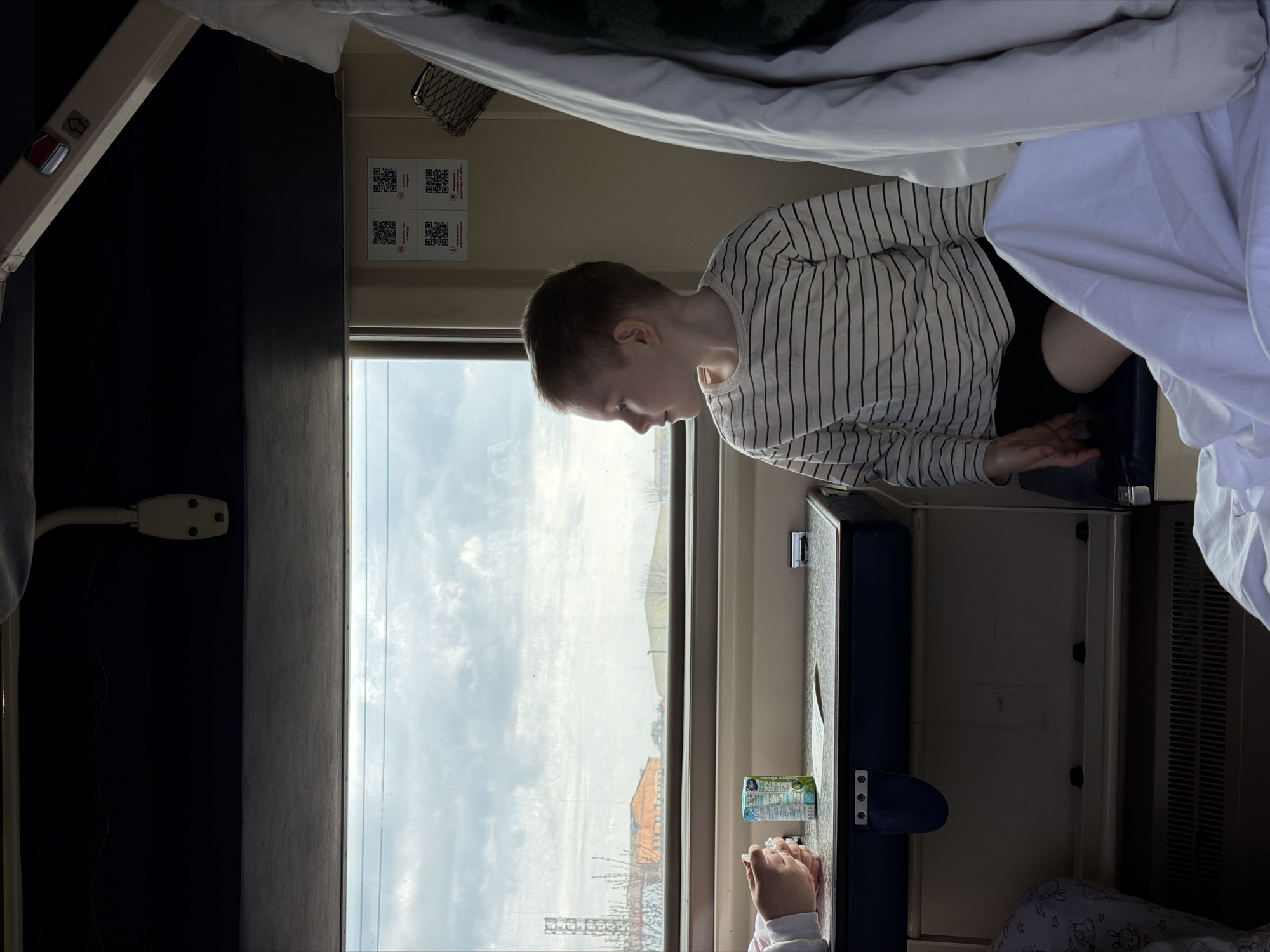 The image depicts the interior of a train compartment. A child wearing a striped shirt sits by the window, looking out. Outside, the scenery suggests a location near Medvyodovskaya, Russia, evidenced by the visible buildings and infrastructure. A second pair of hands rests on the table near a beverage container. The compartment has bunk beds, with white bedding partially visible.