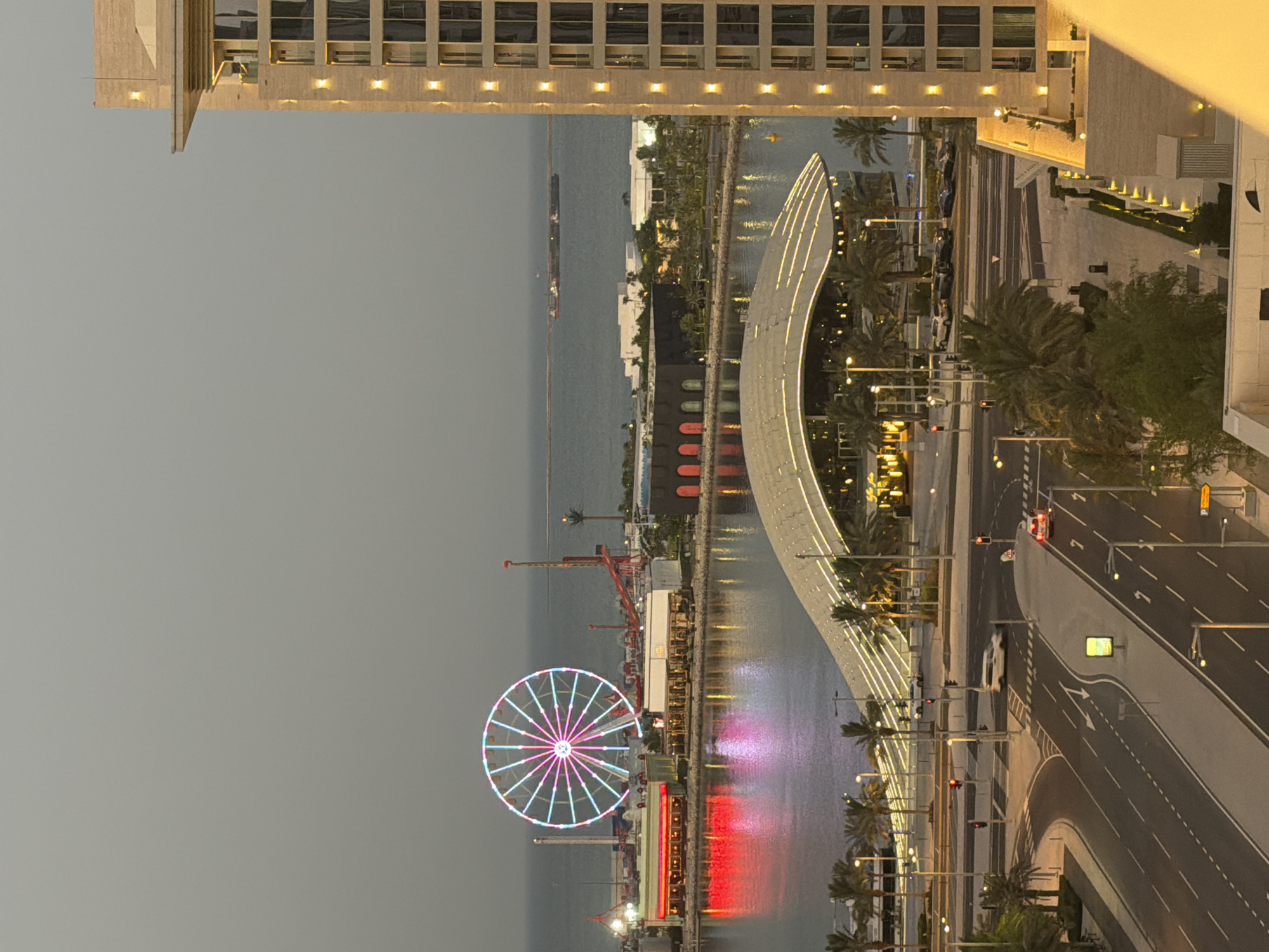 An aerial, eye-level, full shot captures a city waterfront at dusk. In the foreground, a tall, beige apartment building stands to the right, its windows illuminated by warm exterior lights. Below, a busy street with multiple lanes of traffic curves into the scene. Cars are visible on the road, including a white minivan in the middle lane and a white sedan in the right lane, both appearing to be moving. Traffic lights are red and green, indicating the flow of vehicles. Palm trees line the street and sidewalk, adding a tropical feel.
Further back, a modern building with a curved, illuminated roof stretches across the middle of the frame. In front of this structure, a body of water reflects the lights of the city. Across the water, a vibrant Ferris wheel, lit with pink, blue, and white lights, dominates the background. Adjacent to the Ferris wheel are various amusement park rides and buildings, some with red and yellow lighting. In the distance, the calm ocean stretches to the horizon, with a large cargo ship or barge visible on the water. The sky is a muted, overcast gray, typical of twilight.