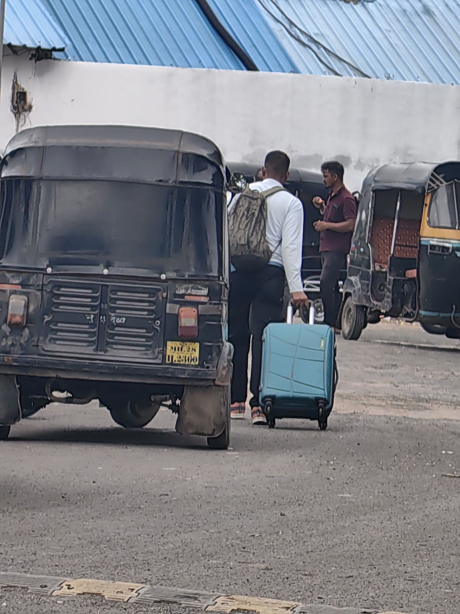 The image depicts an outdoor scene in Malkapur, India, likely a transport hub or roadside. A man wearing a white shirt and a backpack is seen from behind, pulling a teal-colored suitcase with four wheels. He is walking on a paved surface. Behind him, two auto-rickshaws are parked. The auto-rickshaw in the foreground is black with a license plate reading "MH-28 D.2300". Another auto-rickshaw, yellow and black, is parked to its right. In the background, another man in a maroon shirt is visible. A white building with a blue corrugated metal roof is present in the upper portion of the image. The overall environment suggests a functional, perhaps somewhat utilitarian, area.