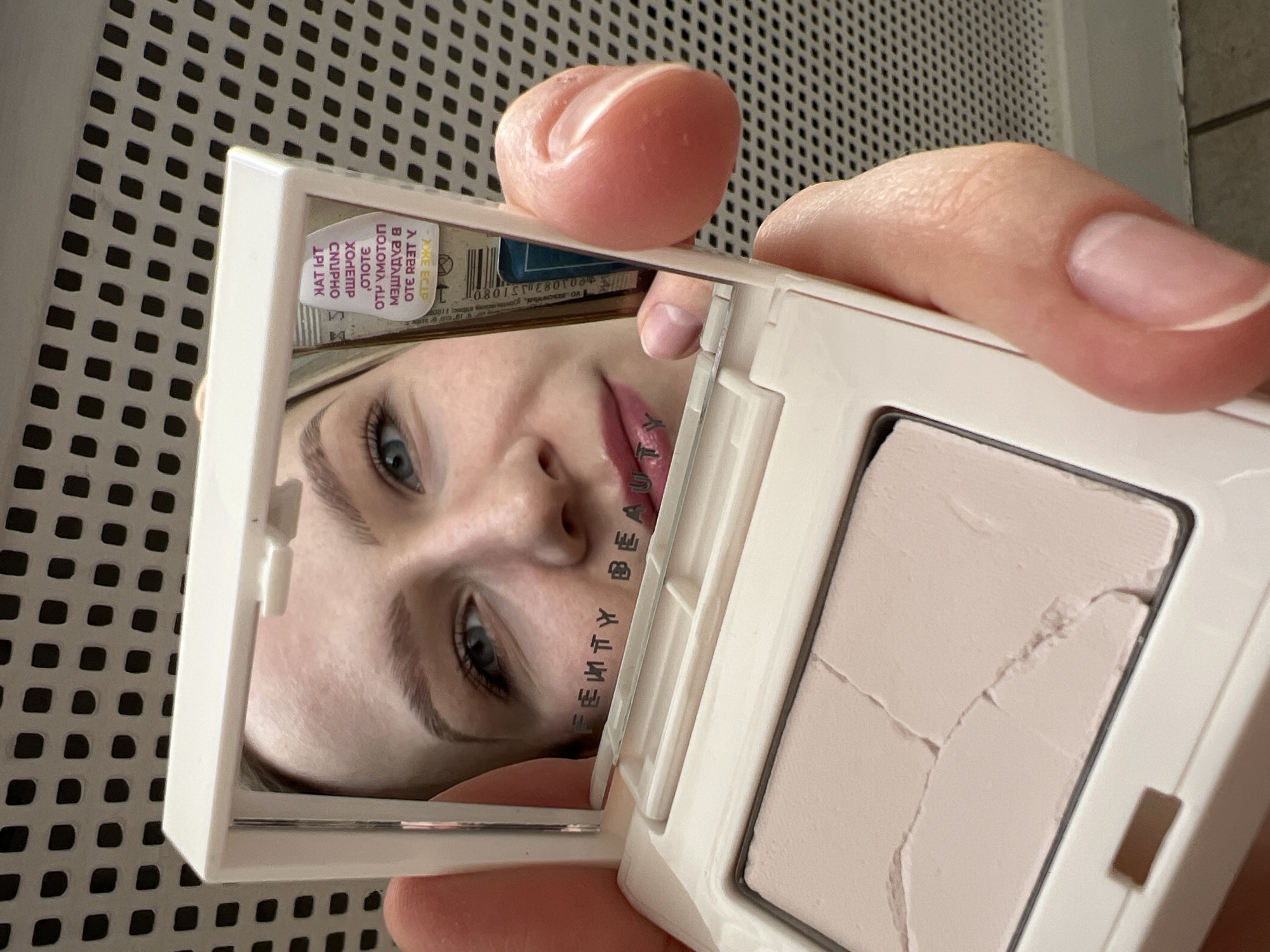 The image shows a woman reflected in the mirror of a white compact powder case. The compact powder itself appears to be cracked. "Fenty Beauty" is tattooed on the woman's face. A label is visible on the mirror containing the city of Khimki, Russia and the address of Kolebsofty, 1120. The background features a white lattice pattern. A person's fingers are visible holding the compact.
