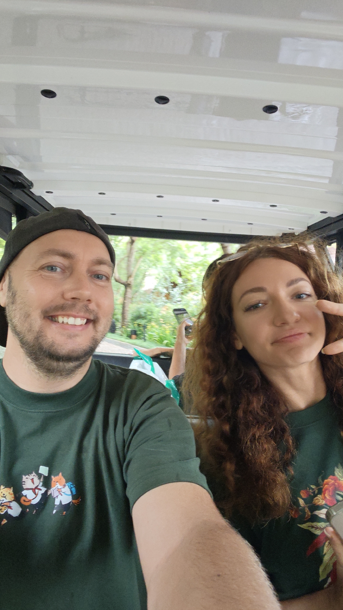 A selfie features two adults, a man and a woman, seated inside an open-air vehicle, likely a golf cart or shuttle, against a lush green background. The man, with light skin, a short beard, and blue eyes, is in the foreground on the left. He wears a dark green t-shirt with a graphic of three cartoon cats dressed in school uniforms and a dark baseball cap worn backward. He smiles widely at the camera.
Beside him on the right, the woman has long, curly red-brown hair, light skin, and sunglasses perched on her head. She wears a dark green t-shirt with a vibrant floral pattern of red, yellow, and green. She makes a playful face and a peace sign with her fingers. The white roof of the vehicle has several small black circular fixtures.
Through the open sides of the vehicle, a dense backdrop of green trees and foliage is visible, indicating an outdoor, natural environment. In the distant background, another person's arm and leg are visible, holding a smartphone. The scene appears to be set during daytime, with bright yet diffused lighting, suggesting either an overcast sky or a shaded area. Both individuals convey cheerful and relaxed emotions. This setting is within the area of Mù Cang Chải, Vietnam.