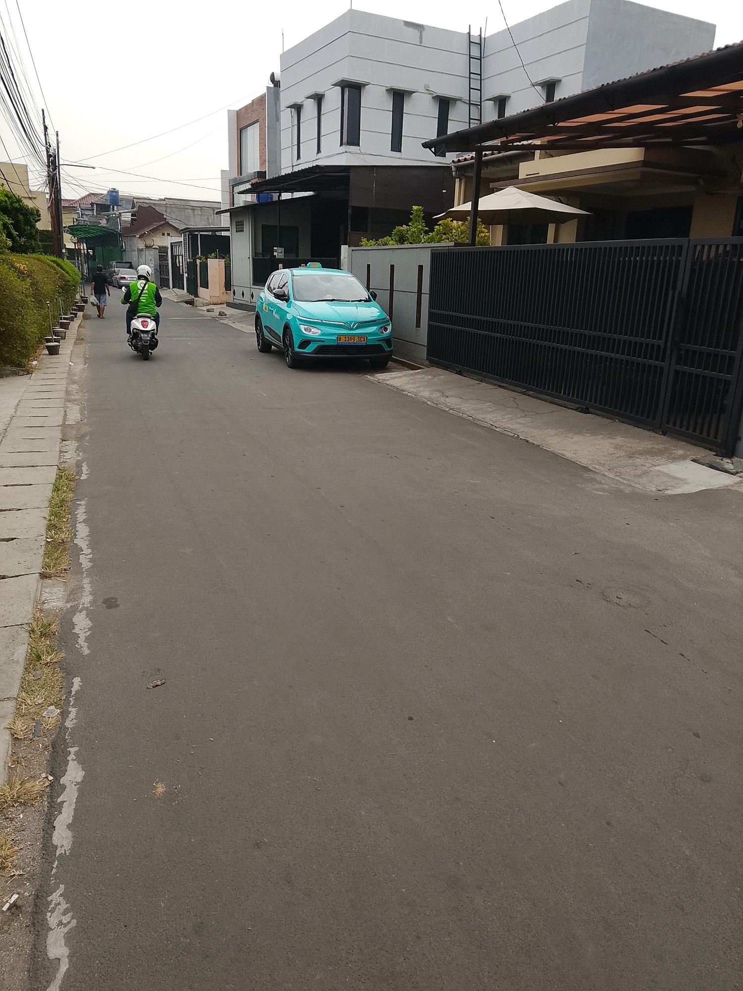 A teal-colored electric car, identified by its license plate "B 1399 SCX," is parked on the right side of an asphalt road in a residential area. A person wearing a green jacket and white helmet is riding a motorcycle on the left side of the road, traveling away from the camera. The road is lined with concrete curbs and a patch of dry grass on the left. On the right, a black metal fence encloses a property with a covered patio area and beige and white buildings in the background. Overhead, power lines are visible against a hazy sky. The scene appears to be in South Tangerang, Indonesia, suggested by the license plate format and architectural style.