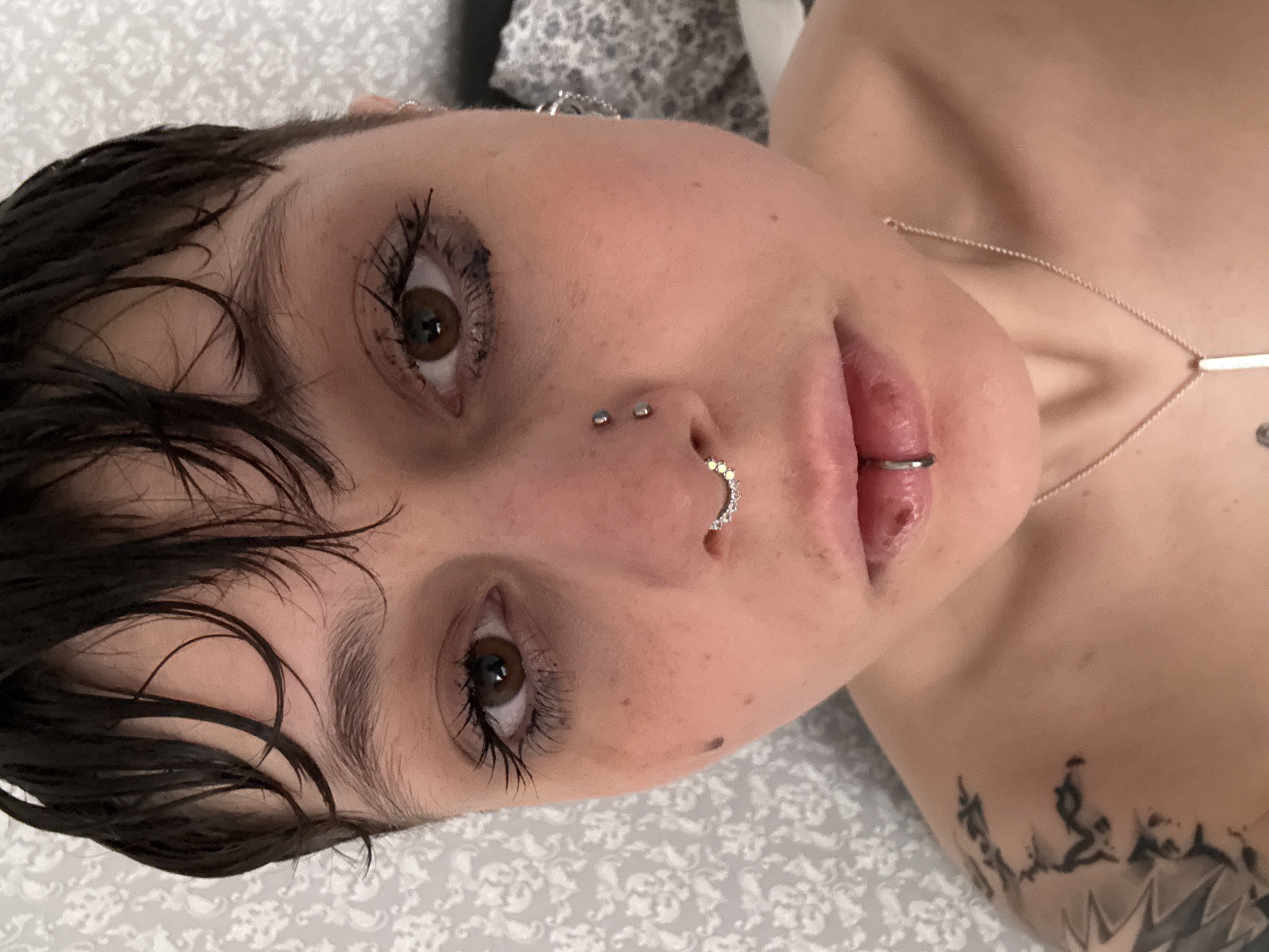 A close-up shot of a person with short, dark, wet hair. The person has brown eyes, multiple nose piercings (two studs above the nostril and a jeweled ring in the nostril), and a lip ring. The skin tone is light. A delicate chain necklace with a bar pendant is visible on the neck. A tattoo is partially visible on the shoulder. The background appears to be a bed with a patterned sheet. The city and country of Saint Petersburg, Russia, are not discernible in this image.