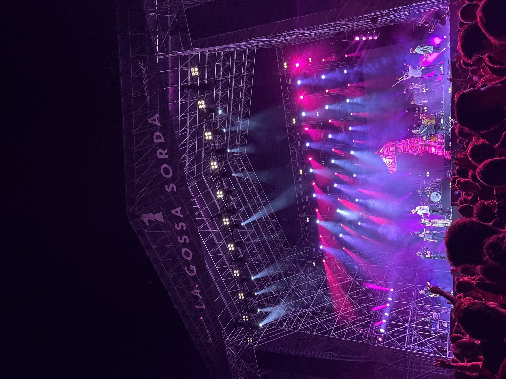 The image shows a concert by the Italian band "La Gossa Sorda" in Cunit, Spain. A large crowd is visible on the right, with their hands raised in the air. The stage is constructed with metal scaffolding and features a complex lighting system using pink and blue lights and lasers. The band members are performing on stage, and a large pink banner is visible behind them. The band's name is printed vertically on the left side of the scaffolding.