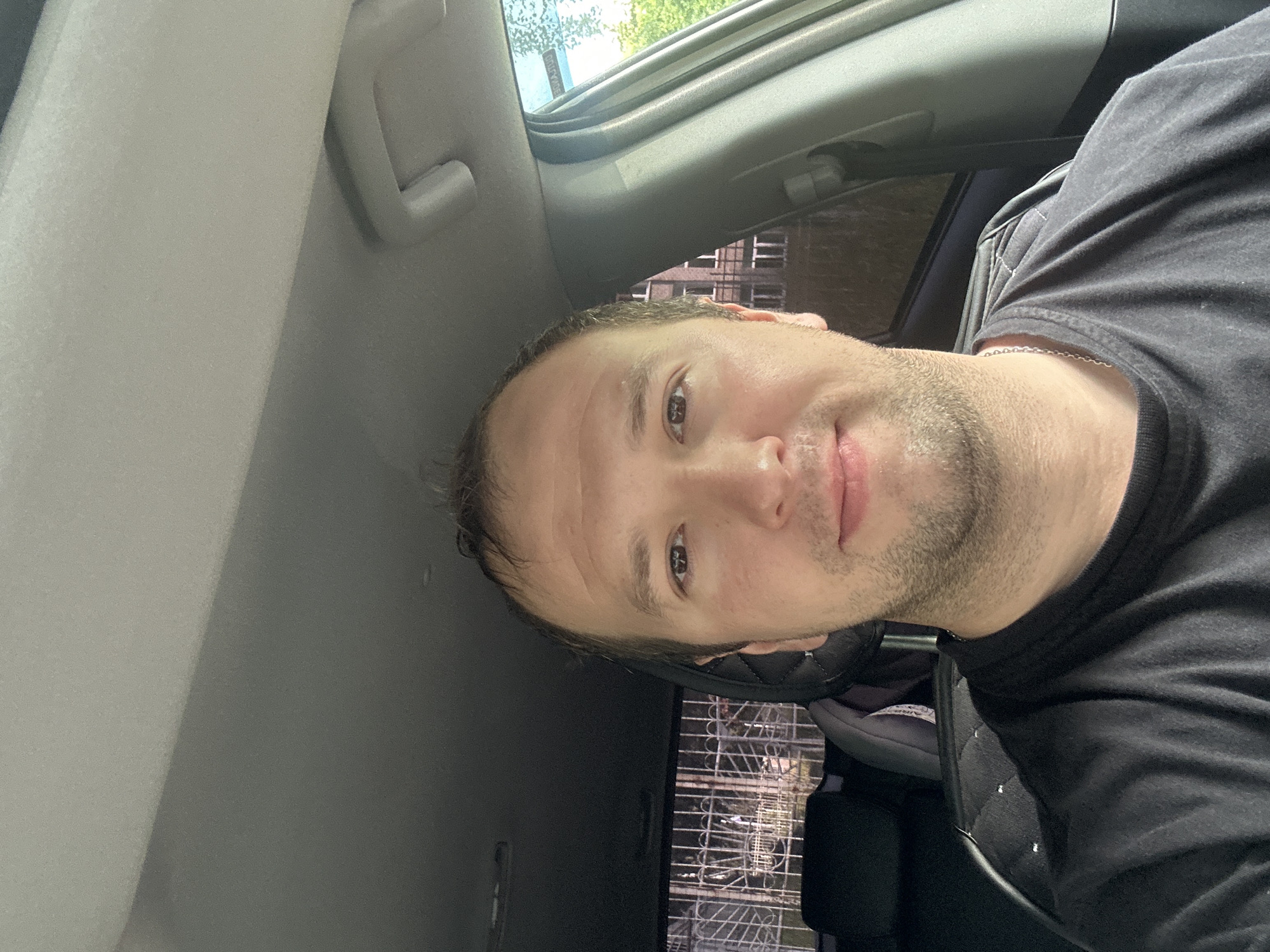 An over-the-shoulder, selfie-style, medium shot shows a fair-skinned man with short brown hair, a light stubble, and brown eyes looking directly at the camera. He is wearing a black t-shirt and a silver chain. He is seated in the driver's seat of a car, with the interior visible. The man's head and shoulders are the primary focus. The background is out of focus but suggests an urban environment. The location is identified as Chaikovsky, Russia.