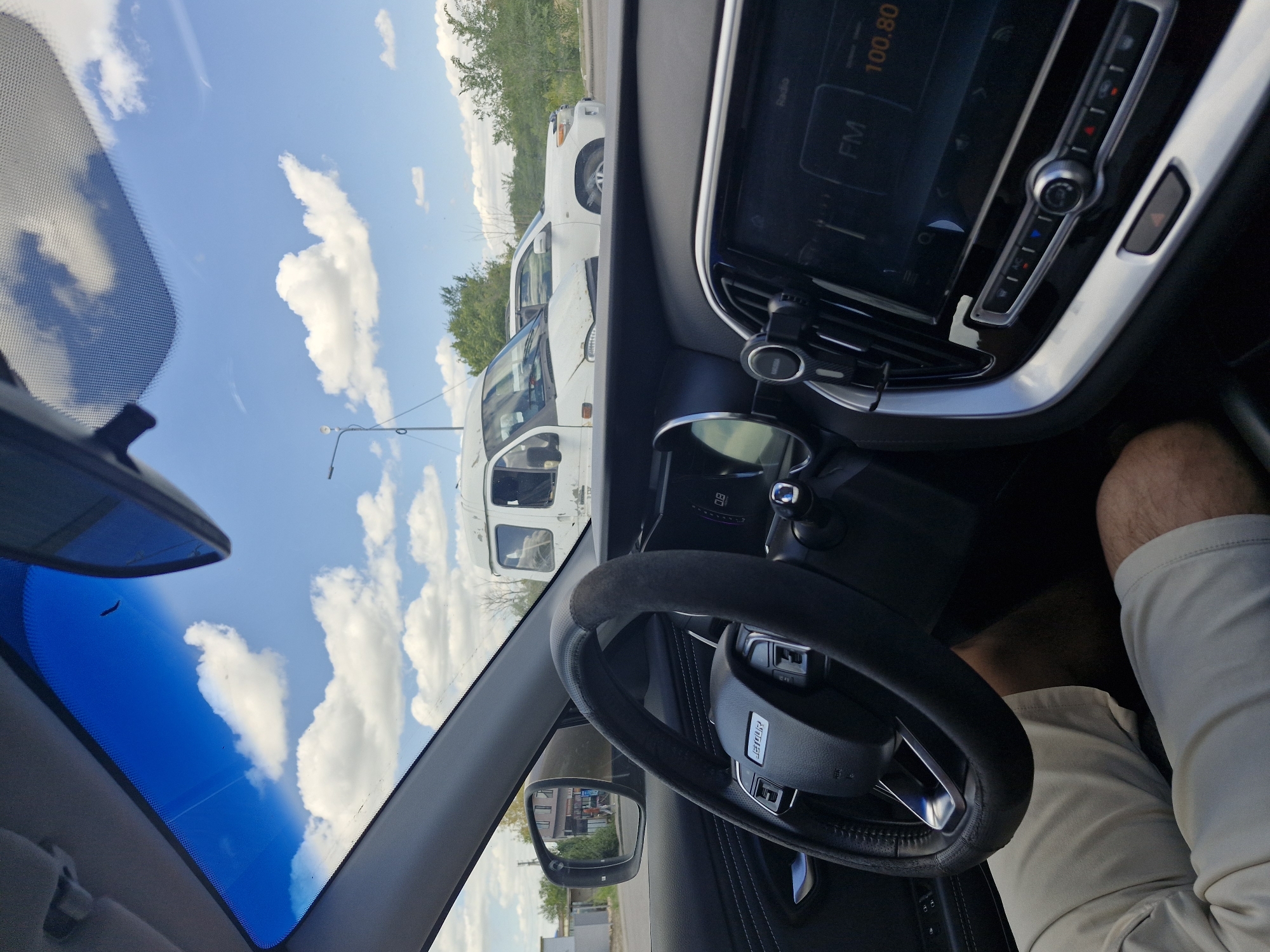 This is a point-of-view shot from inside a car, looking out the windshield and towards the dashboard and steering wheel. Outside, the sky is blue with fluffy white clouds. There are two white vehicles visible on the road in front of the car, a van and what appears to be an SUV. Trees and a building are visible in the background. The dashboard features a digital display showing "FM 100.80" and a steering wheel with a leather cover. A person's legs in light-colored shorts are visible in the lower part of the frame. The image appears to have been taken in Astana, Kazakhstan.