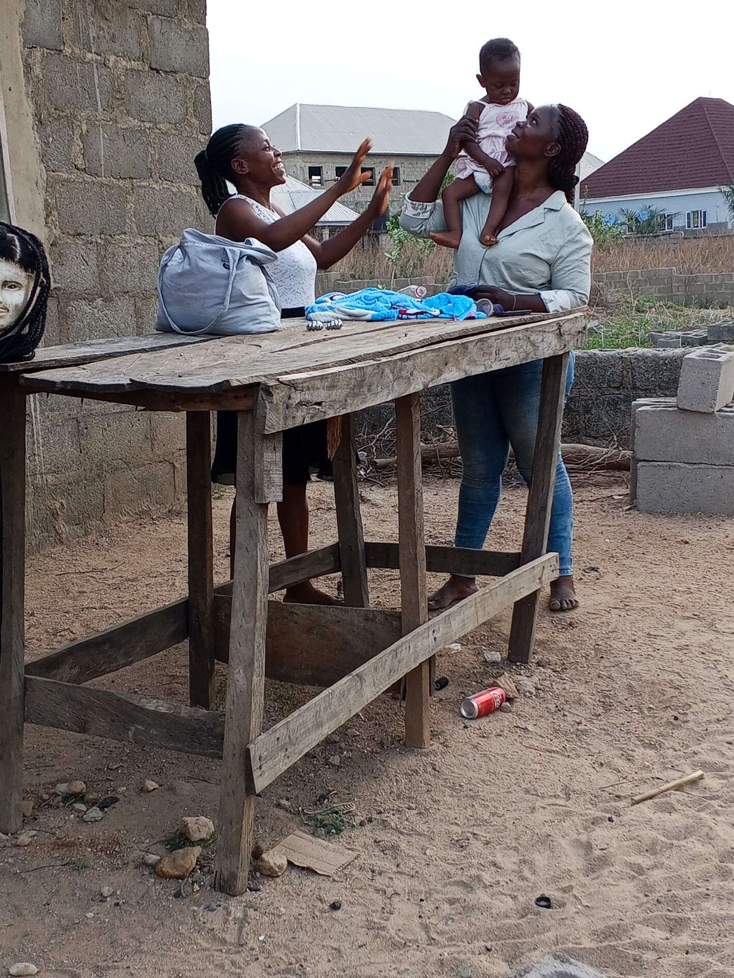 The image depicts two women interacting near a wooden table in an outdoor setting. One woman, wearing a white top, has her arms raised and appears to be smiling or laughing. She is positioned next to the table, which has a light-colored bag, a blue towel and other unidentifiable objects on it. The other woman is holding a baby on her shoulder. She is wearing a light-colored shirt and jeans, and is barefoot. In the background, there's a partially constructed brick wall and a residential building. The ground is sandy and littered with a red can and other debris. The scene appears to be in a developing area, possibly in the city of Adonkolo, Nigeria, as indicated in the prompt. Additional cinder blocks are visible in the background.