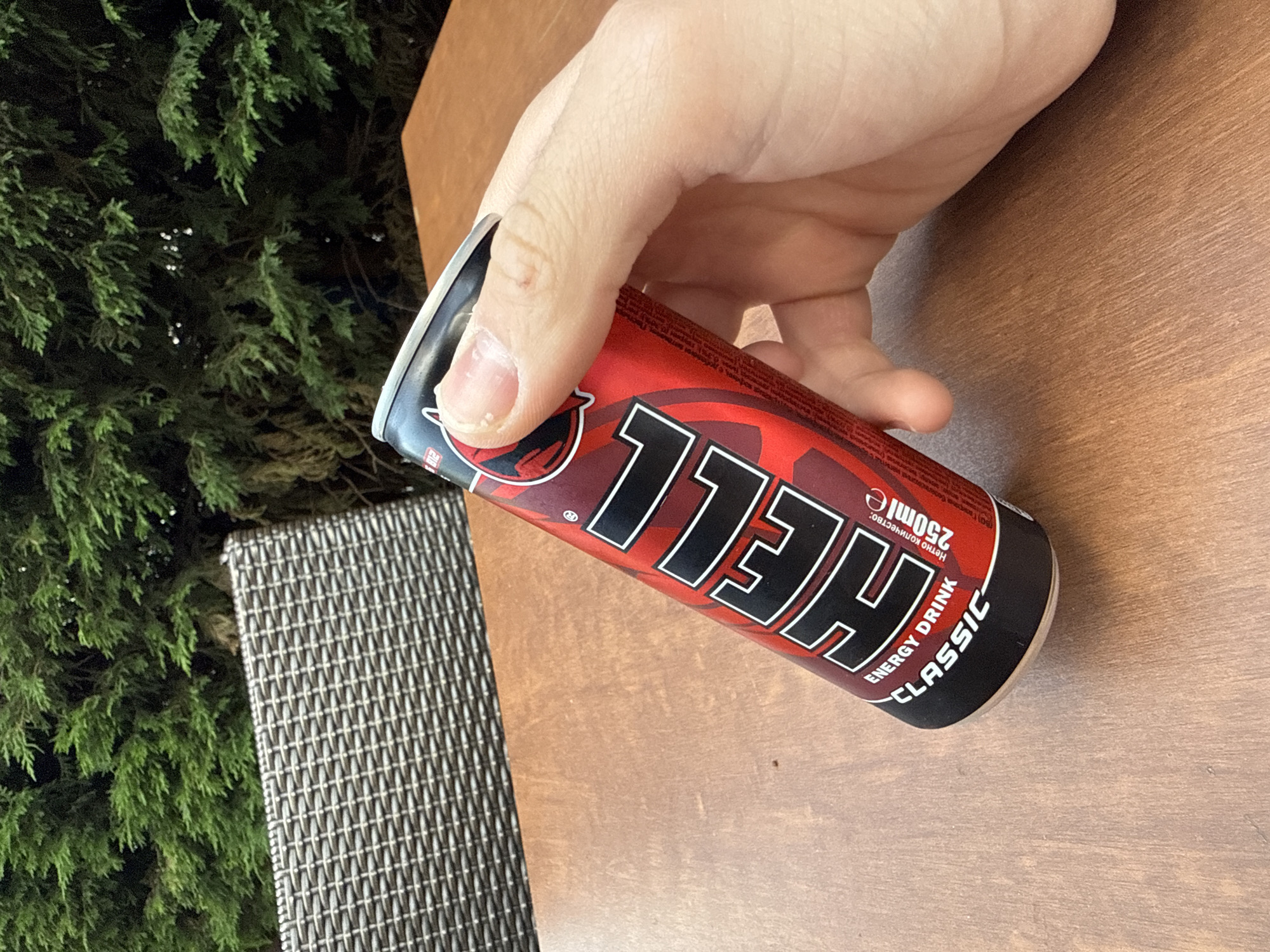 A hand is holding a red and black "HELL" energy drink can on a wooden surface. Part of a woven chair is visible to the left. In the background is a green shrub. The brand name "HELL" is written on the can. The text "Energy Drink Classic" and "250ml" are visible on the can. There is no explicit indication of Stara Zagora, Bulgaria.