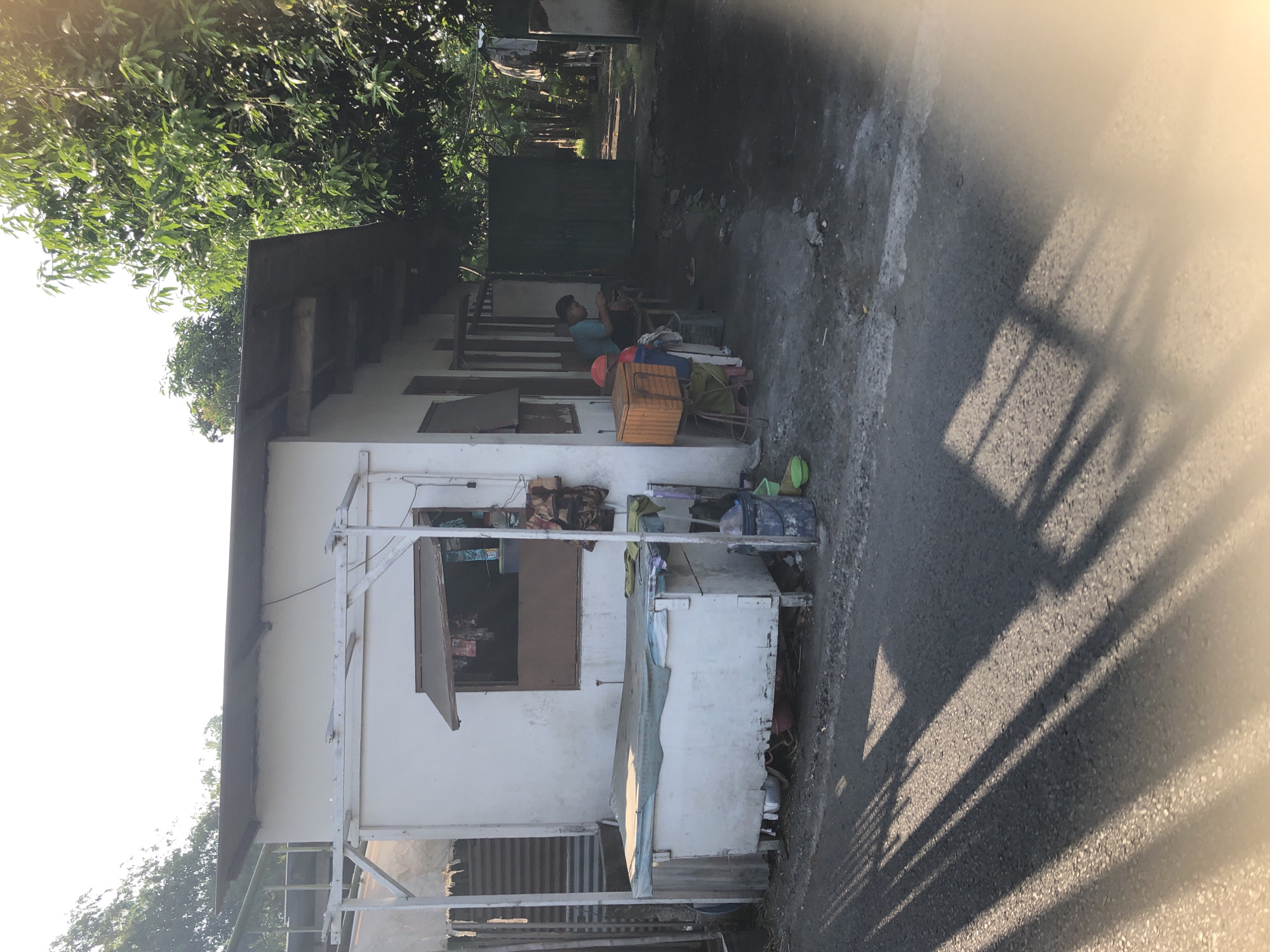 The image depicts a small, white, single-story building with a slanted, dark-colored roof. The building appears to be a shop or kiosk. In front of the building, on the left side, is a long, white chest freezer covered with a green tarp. To the right of the freezer, a small window with a brown awning is visible, displaying various items for sale. Next to the window, on the exterior wall, a patterned cloth is hanging.
Further to the right, a man in a blue shirt is seated on a stool near an open doorway. In front of the man, there is an orange crate and other items. The ground in front of the building is unpaved and appears wet in places, with some debris scattered around. A large tree with dense green foliage is visible behind and to the right of the building, casting shadows across the scene. The shadows of what appear to be the building's structure and possibly nearby trees are cast onto the asphalt road in the foreground. The overall impression is a humble, roadside establishment in a tropical environment, possibly located in Putat, Indonesia, given the vegetation and architecture.