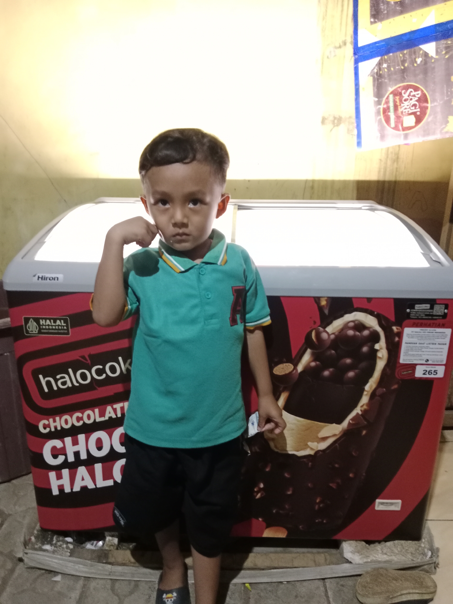 A young boy stands in front of a frozen food display freezer in what appears to be an outdoor or semi-enclosed area. The freezer is branded with "halocok" and features an image of a chocolate ice cream bar. The boy is wearing a teal polo shirt and black shorts. He has his left hand on his cheek in a thoughtful pose. In the background, a wall is visible with a yellow light illuminating the scene. A sign with "Hiron" is affixed to the freezer. A sticker on the right side of the freezer shows the number "265" in a red rectangle. The ground beneath the freezer is made of rough stone and cement. The location is identified as Parage, Indonesia.