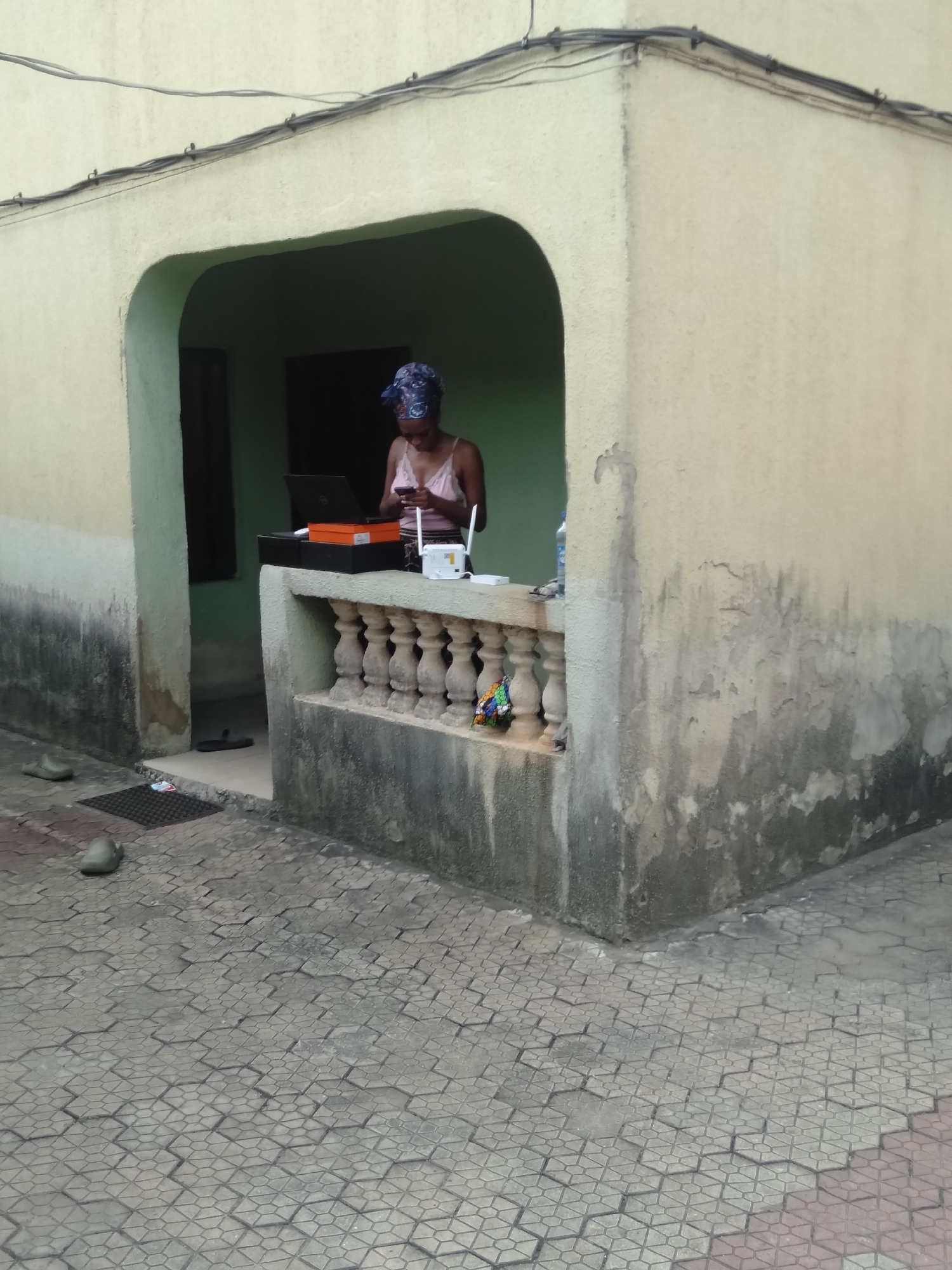 A young woman stands under a semi-enclosed porch or entryway of a building in Shangisha, Nigeria. She is wearing a light pink tank top and a patterned blue and white headwrap, focused intently on a smartphone held in both hands. Her head is bowed as she interacts with the device.
To her left, on a low balustrade or railing, an open black laptop rests on top of an orange box and some other dark boxes. Next to the laptop, a white internet router-like device with two prominent antennas and a visible "1" on its front is placed. A small, colorful, possibly crocheted or woven item is tucked into the balustrade below the router.
The building's exterior walls are painted a pale yellow or cream, but show significant signs of wear, including dark staining, discoloration, and peeling paint, particularly on the lower half. Overhead, several utility wires are strung across the facade, secured by small hooks. The ground in front of the porch is paved with interlocking grey and dark grey hexagonal and rectangular blocks, forming a geometric pattern. A black doormat and a pair of worn, light green clogs are visible on the paved area near the entrance. The entrance itself is a dark opening, suggesting a room beyond.
The scene depicts a moment of connectivity, possibly work or leisure, in an everyday setting. The natural lighting suggests it is daytime, though the specific time is not clear.