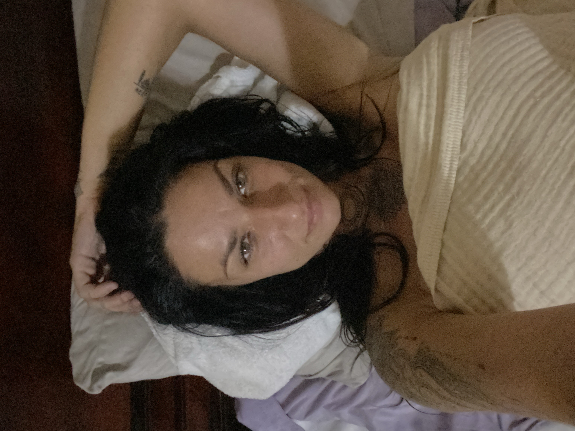 A woman with dark hair and light eyes is depicted lying on a bed in a low-light indoor setting in Mũi Né, Vietnam. She is positioned on her back, looking directly at the camera with a neutral expression. Her hair, appearing damp or wet, spreads across a white pillow. Her right arm (viewer's left) is bent with her hand near her hair, while her left arm (viewer's right) is extended upwards, with her hand behind her head, out of frame. She is wrapped in a light-colored, possibly cream or beige, ribbed towel or blanket that covers her torso. Visible tattoos include a large, intricate design on her left upper arm/shoulder (viewer's left), a circular pattern on her upper chest, and a small, dark, abstract design on her right inner forearm (viewer's right). The bed features white and light purple bedding, and a dark wooden headboard is visible in the background.