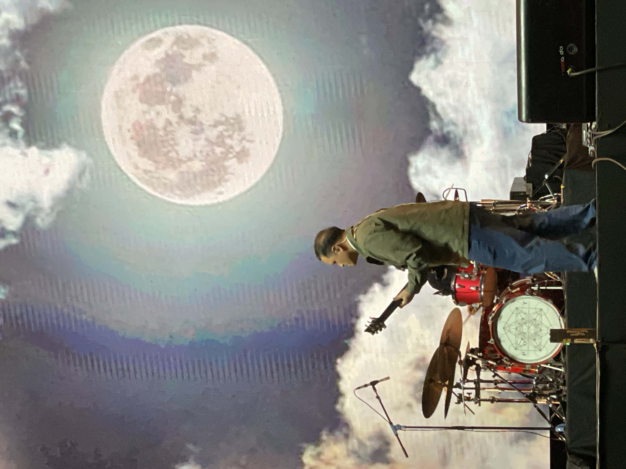 A musician plays guitar in front of a drum kit on a stage, set against a backdrop featuring a large moon and cloudy sky. The drum kit is red and silver, with a design on the front drum. The musician is wearing a green jacket and blue jeans. The location is likely Salembaran, Indonesia, based on the user's request.