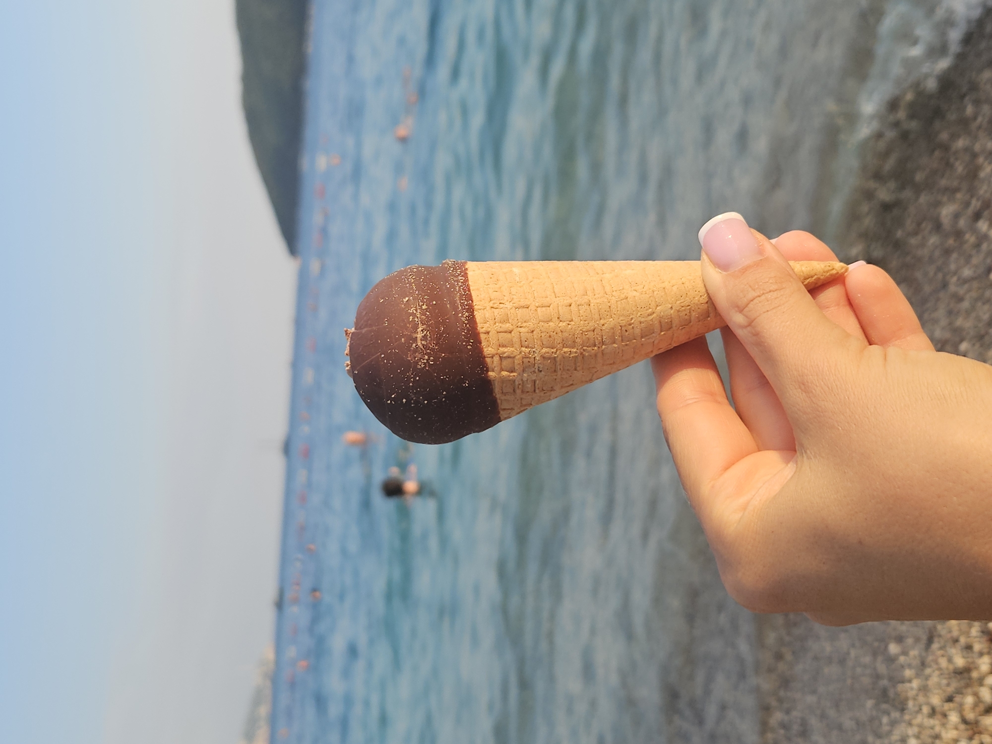 A close-up shot features a hand holding an ice cream cone topped with chocolate in front of a blurred beach scene. The hand, with a French manicure, gently grips the waffle cone. In the background, the turquoise sea is visible, with buoys marking a swimming area and several out-of-focus figures in the water. A hilly coastline stretches across the horizon under a clear sky.