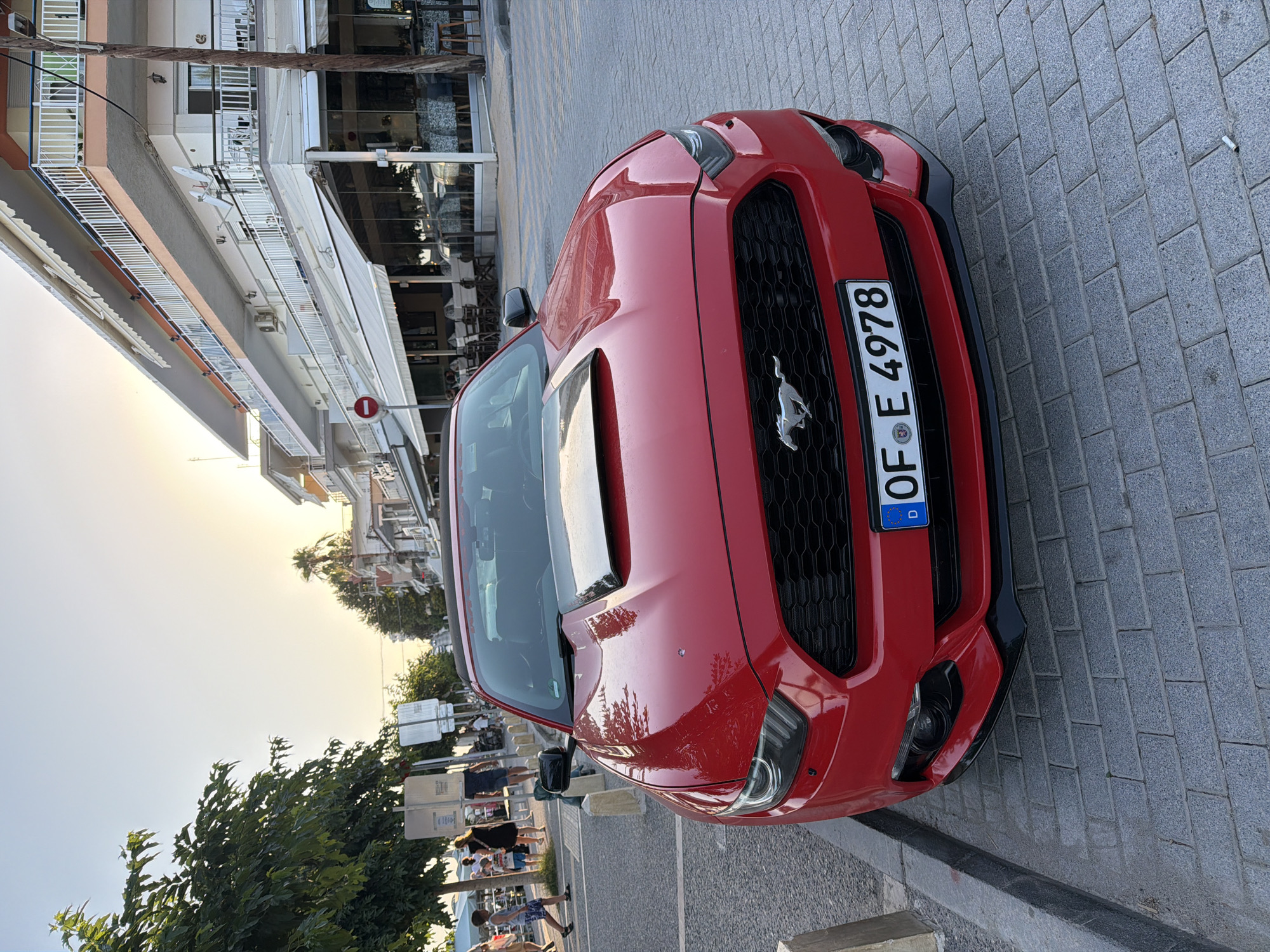 A red Ford Mustang is parked on a sidewalk next to a street with cobblestone paving. The car is facing the viewer's right and its license plate is visible, reading "OF E 4978" with a European Union emblem. The car has a black grille with a silver Mustang logo. In the background, there is a building with shops and restaurants, and trees. The sky is bright, suggesting it is daytime, likely late afternoon or early evening given the warm lighting. There are people walking on the sidewalk in the background, some appearing to be tourists. The scene appears to be in a Mediterranean coastal town. A red and white circular "no entry" sign is visible on the building in the background. The overall impression is a snapshot of a car in a pleasant, public setting during a bright day.