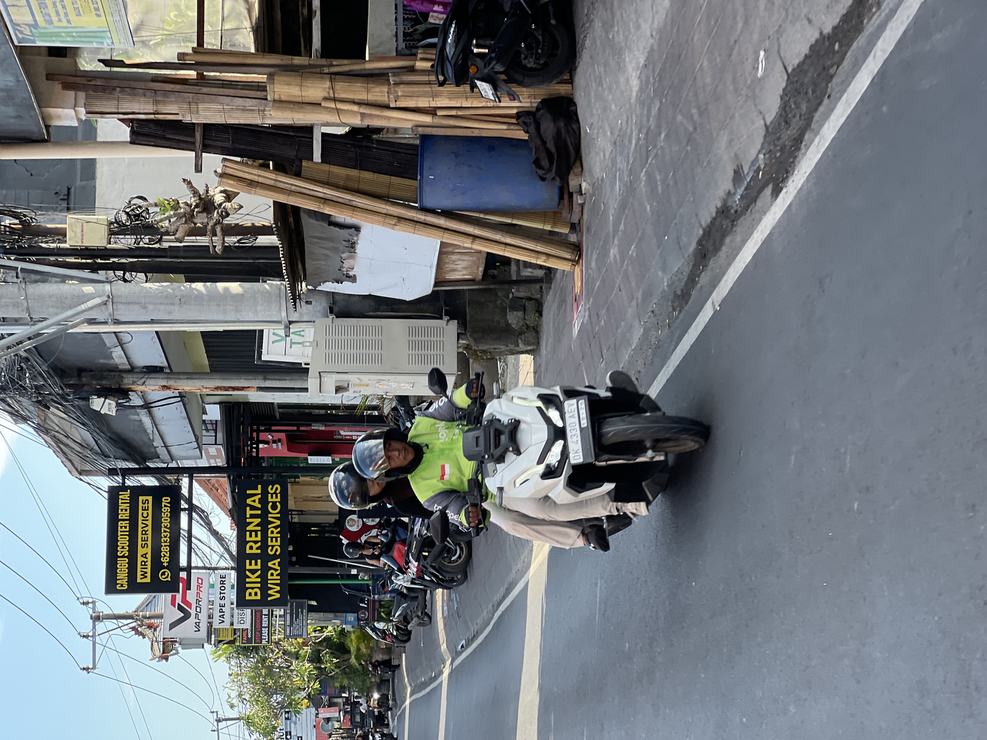A white motorcycle carries two individuals as it travels down a street in North Kuta, Indonesia. The driver, a male wearing a neon green jacket and a black helmet, operates the vehicle. Behind him, a female passenger also wears a black helmet. The motorcycle's license plate reads "DK 4330 AEV". The street is paved and bordered by a sidewalk on the right. The sidewalk has several bamboo poles stacked vertically, a large blue plastic barrel, and a dark cloth bag on the ground. Buildings with signs are visible along the street, including one that says "CANGGU SCOOTER RENTAL WIRA SERVICES" and another with "BIKE RENTAL WIRA SERVICES". Overhead, a complex network of electrical wires is strung between poles. The scene is illuminated by natural daylight, suggesting it is daytime.