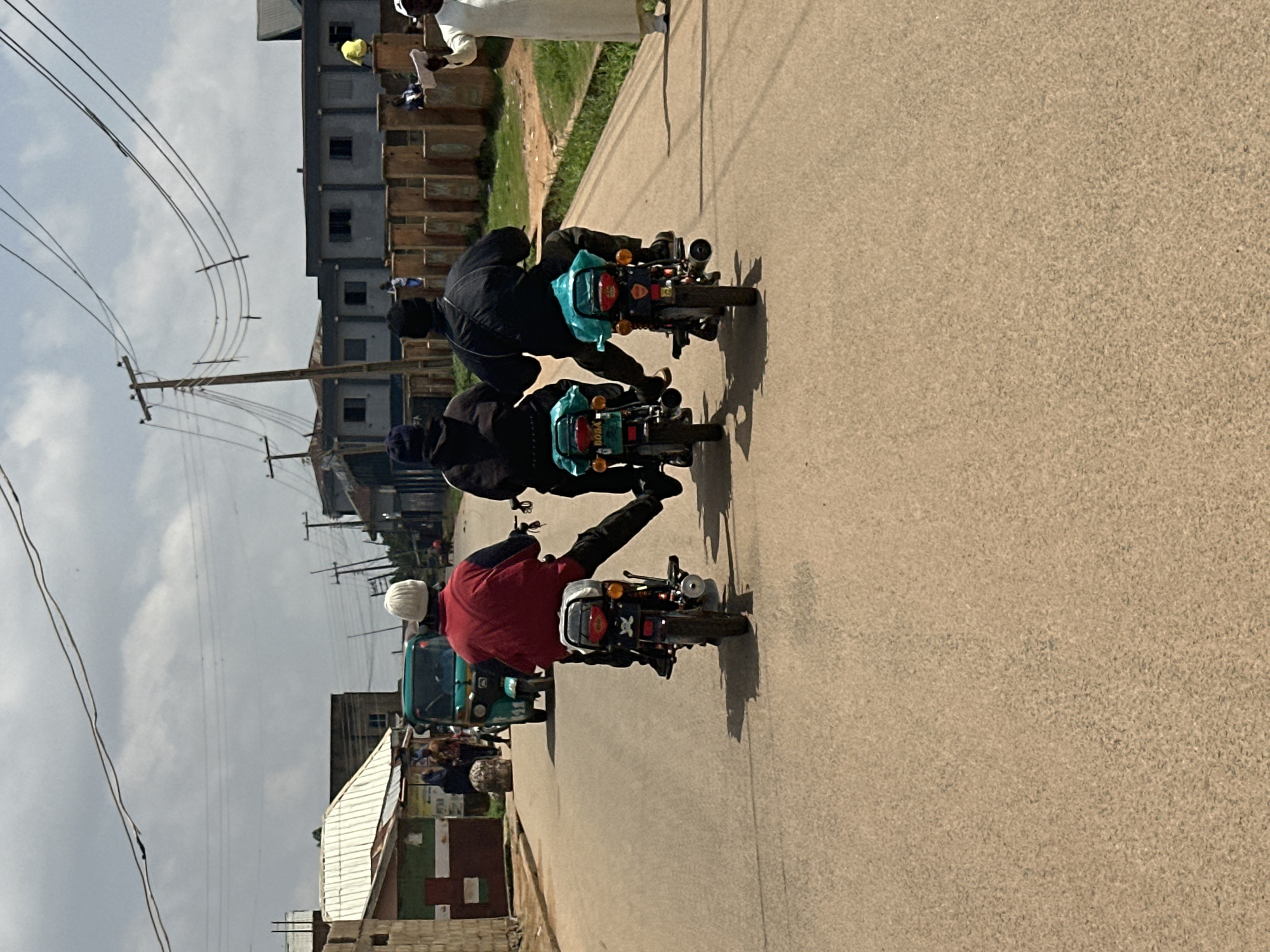 In Jos, Nigeria, three individuals are seen riding motorcycles in a single file on a paved road under a partly cloudy sky. The road is lined with buildings, some appearing to be shops or residential structures, and utility poles with numerous wires. The first motorcyclist, in a red jacket and white beanie, is positioned on a black motorcycle. Following closely are two individuals on dark-colored motorcycles, one with a turquoise tarp on the back. On the right side of the road, a man in a white robe and cap is holding a piece of paper and appears to be observing or directing. A teal-colored auto-rickshaw is visible further up the road, in front of a small shop. The scene depicts a typical street scene in a Nigerian city.