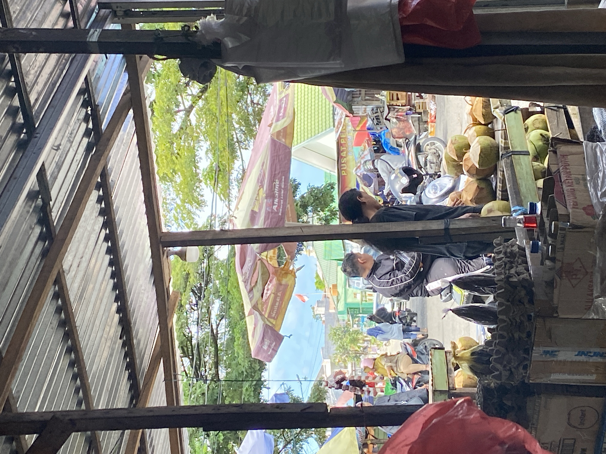 The image captures a street market scene in Ta', Indonesia. The foreground is dominated by a stall featuring numerous coconuts, some piled on wooden planks and others in cardboard egg cartons. Boxes are visible below the coconuts. To the left, a red plastic bag hangs.
In the midground, two men are present. One is wearing a black tracksuit with white stripes and is seated on a motorcycle. The other man, wearing a black long-sleeved shirt, stands near him. Behind them, several other motorcycles and people are visible, some riding and some walking. An orange and pink striped umbrella is open above the market stalls, displaying the "Alkaline" logo. Further in the background, buildings with green roofs and a white spire are visible, along with trees and a bright blue sky with a flag. The market appears to be situated along a street with vehicle traffic. The overall environment suggests a bustling outdoor market on a sunny day.