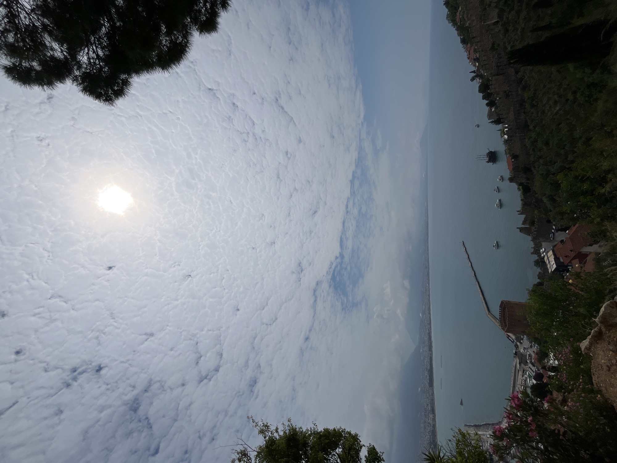 This image captures a high-angle view of Alanya, Turkey, overlooking the sea. The foreground is dominated by foliage, including trees and flowering plants. The middle ground features a long pier extending into the water, with several boats visible near the shore. The city of Alanya stretches along the coastline in the background, partially obscured by haze or clouds. The sky is filled with a layer of clouds, and the sun shines brightly.