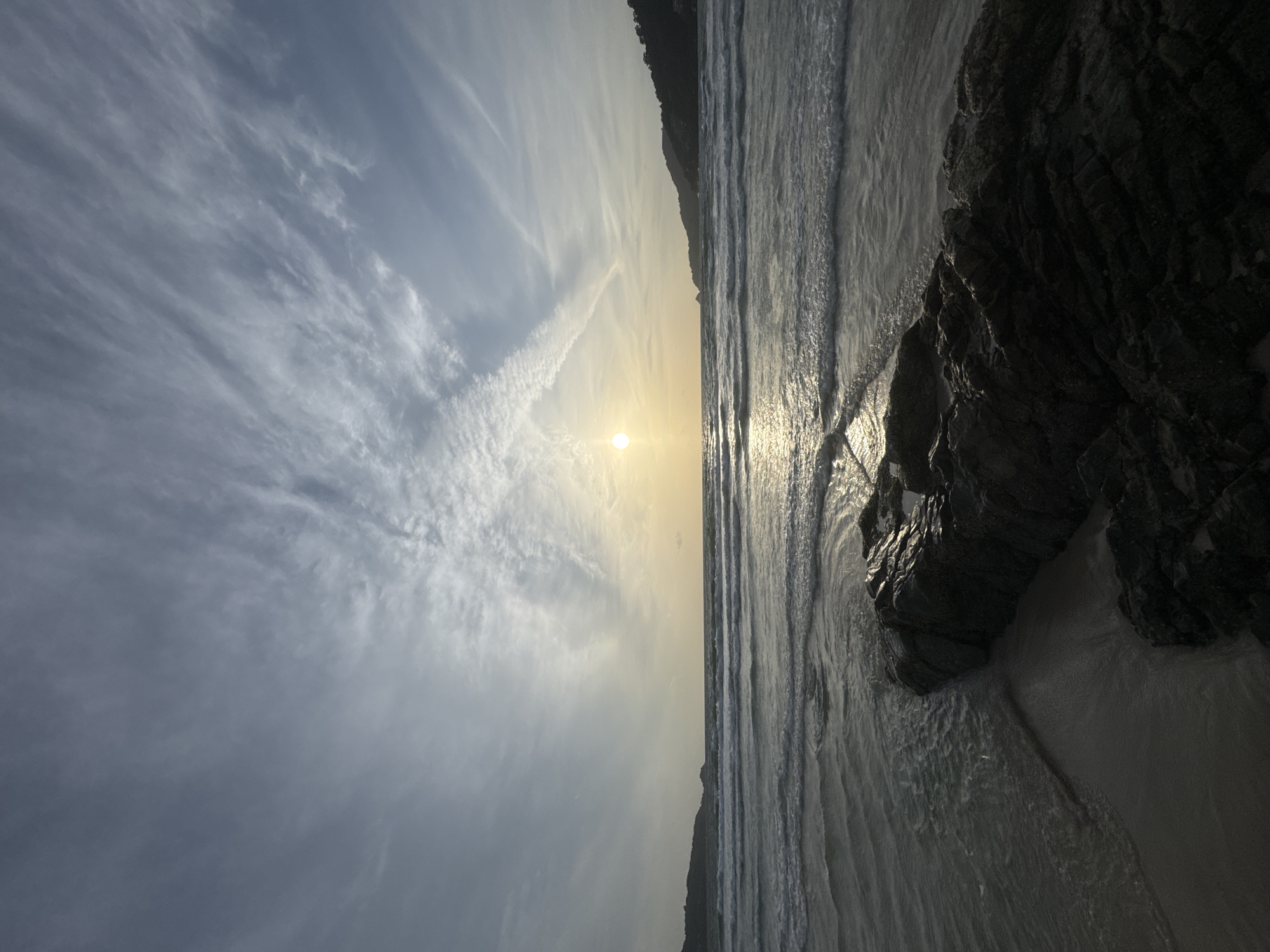 The image depicts a serene beach scene in Patong, Thailand, likely during sunrise or sunset. The sun is a bright, warm orb low on the horizon, casting a golden shimmer across the gentle waves of the Andaman Sea. Wispy, white clouds streak across the pale blue sky, forming an interesting pattern. In the foreground, a dark, rugged rock formation emerges from the wet sand, with small waves lapping at its base. The coastline curves gently in the distance on either side, with lush green hills rising behind the beach, hinting at the tropical landscape of Patong. There are no people visible in the frame. The overall atmosphere is tranquil and natural.