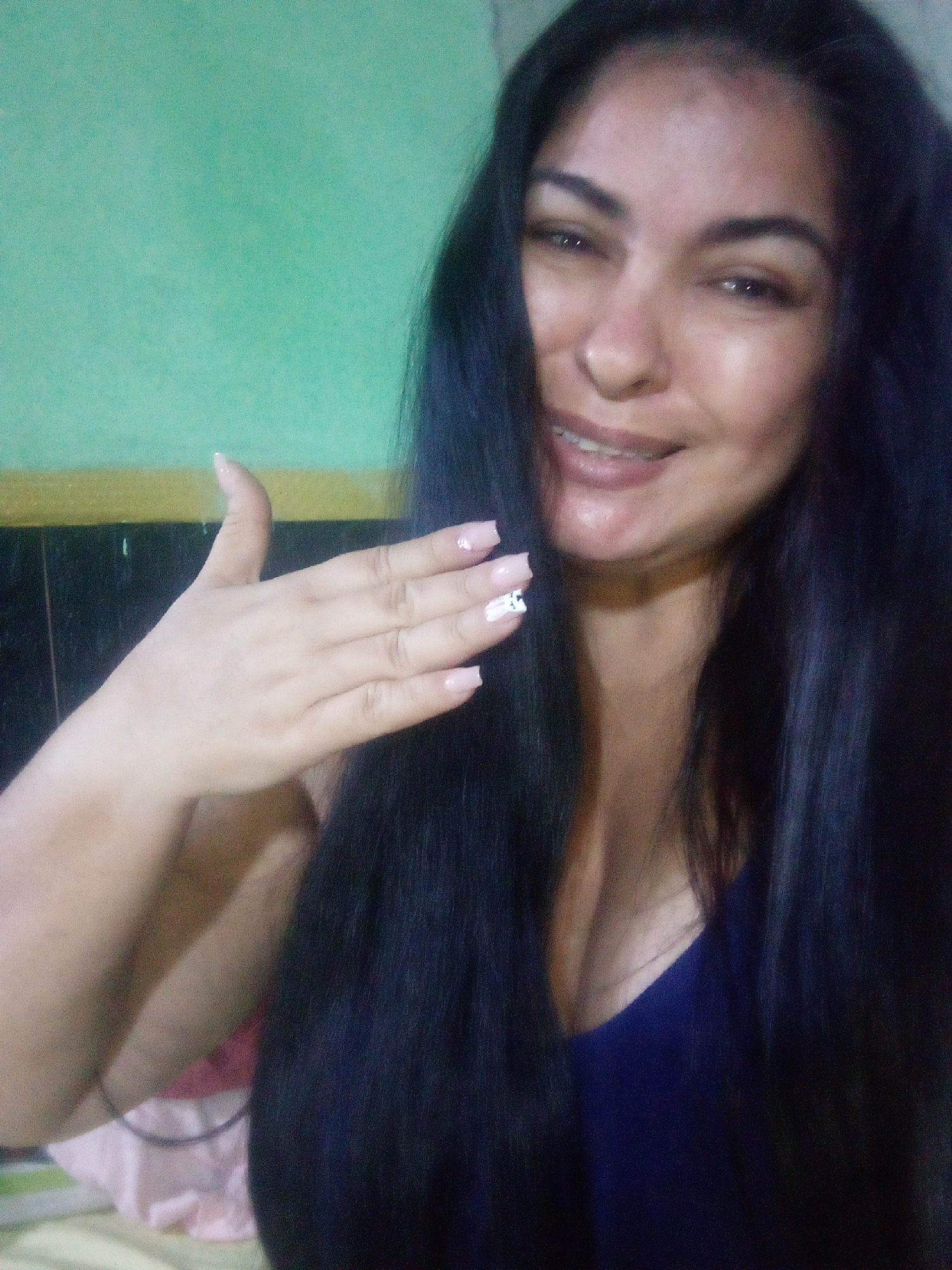 A woman with long, dark hair is smiling and holding up her left hand, which has pale pink nails with white floral designs on some fingers. She is wearing a navy blue top. The background consists of a green wall, a black object, and a glimpse of a bed or couch with a pink and white patterned fabric. The location is unknown.