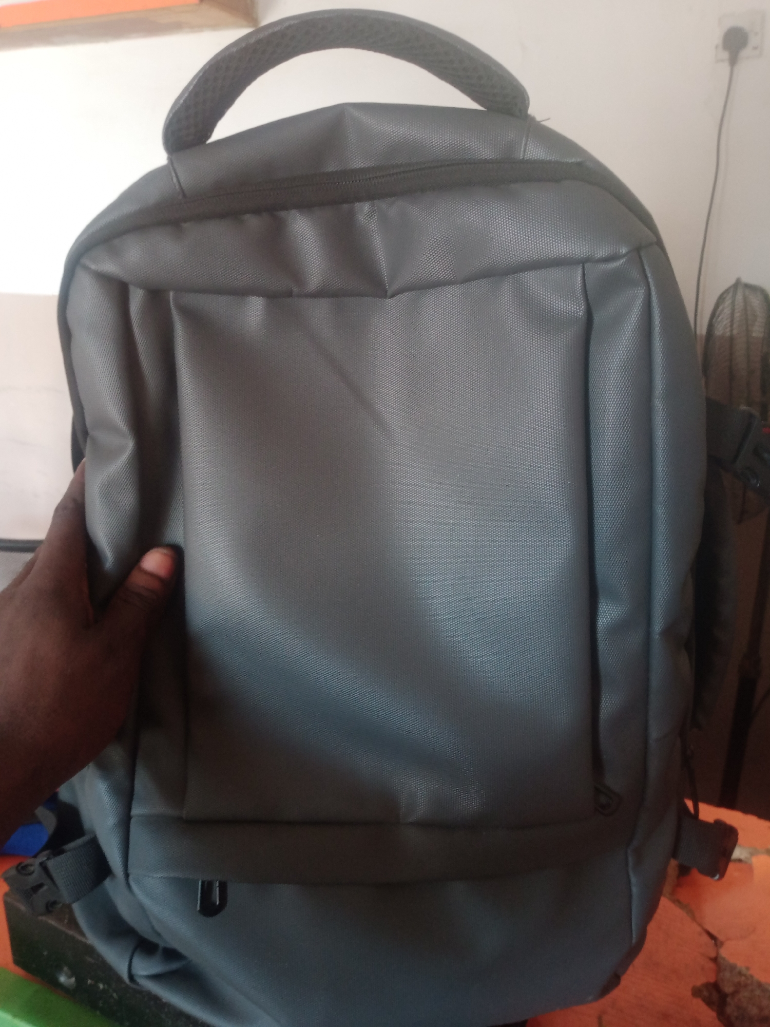 The image shows a close-up of a gray backpack. The backpack is made of a textured material and has a handle at the top. A person's hand, with dark skin, is visible on the left side of the frame, appearing to hold or support the backpack. The backpack is positioned in what appears to be an indoor setting, with a blurred background that includes a white wall, an electrical outlet with a plug, and a fan. The overall lighting suggests an ambient indoor environment. The location is identified as Jalingo, Nigeria, although no specific urban features are visible in the image itself to confirm this.