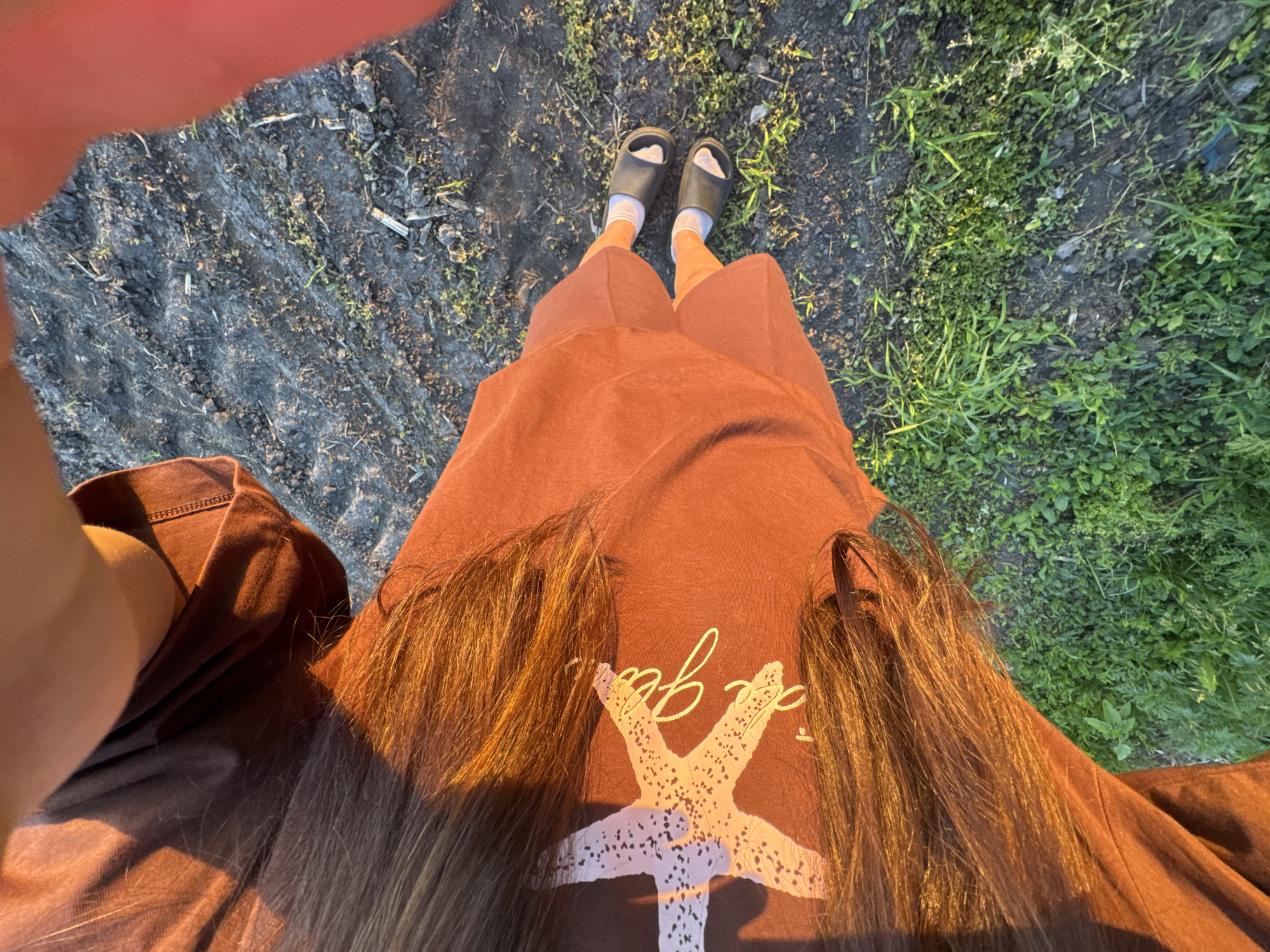 A high-angle forensic perspective captures an individual standing outdoors in Ohuivka, Ukraine. The subject wears a rust-colored t-shirt adorned with a white starfish graphic and partially legible script, layered over matching rust-colored shorts. Long, dark brown hair cascades over the shoulders and chest, illuminated by direct sunlight which also casts warm tones on the attire and creates shadows. White socks and black slide sandals are visible on the feet. The ground below is a mix of verdant green vegetation on the left and darker, disturbed earth with sparse growth and scattered debris on the right, suggesting an agricultural or garden setting. The individual appears to be taking a self-portrait, evidenced by the perspective and the partial view of an arm holding a device on the right side of the frame.