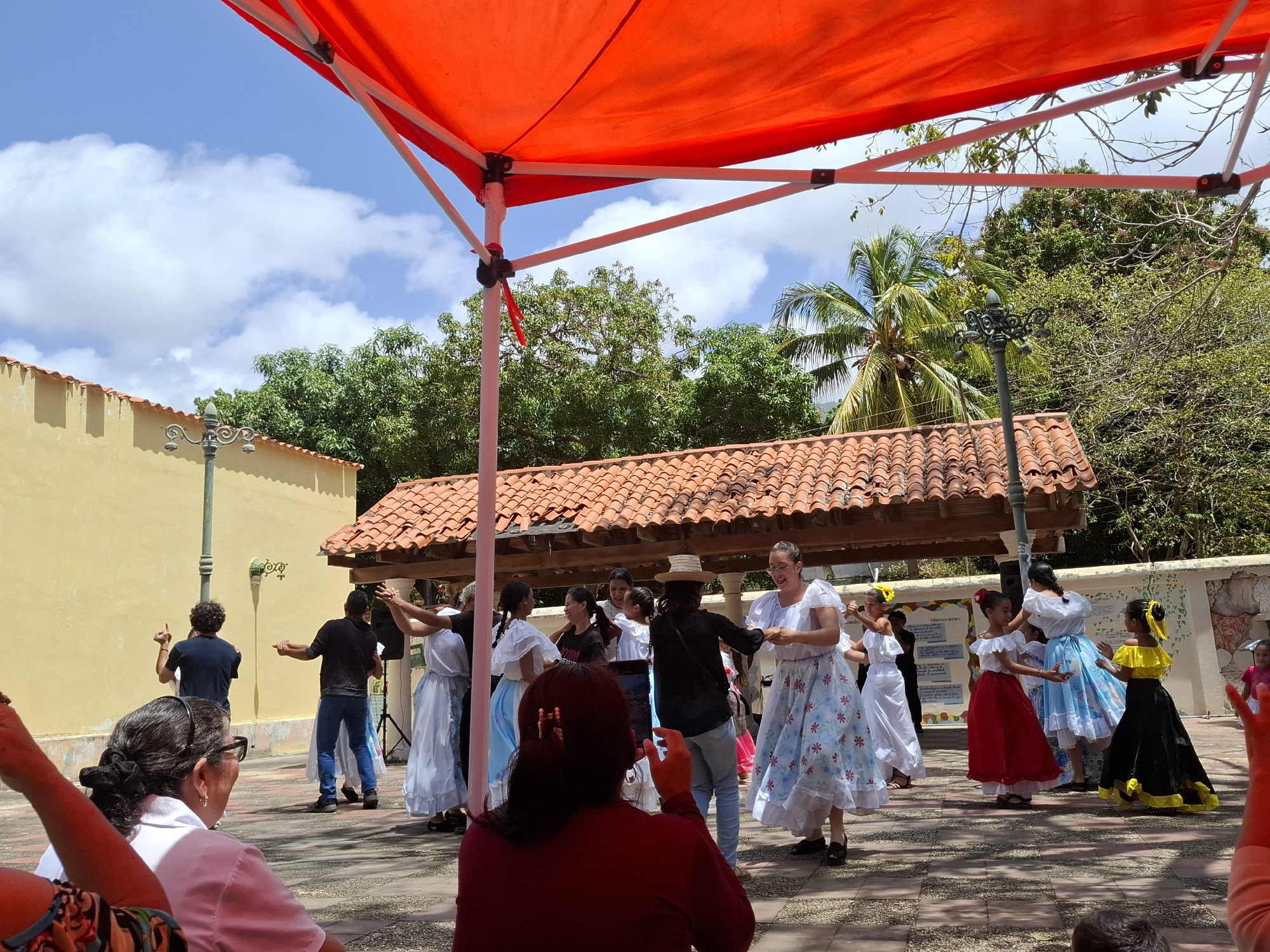 A group of people is dancing in El Valle del Espíritu Santo, Venezuela. A partially visible red awning is in the foreground, above a crowd of people who appear to be watching or filming the dancers. Among the dancers, both adults and children are seen, with several of them dressed in traditional attire. They are actively engaged in a dance performance. Some onlookers have their arms raised, perhaps taking photos or cheering. The background features buildings with red tile roofs, lush trees, and a partly cloudy sky. A decorative street lamp is also visible.