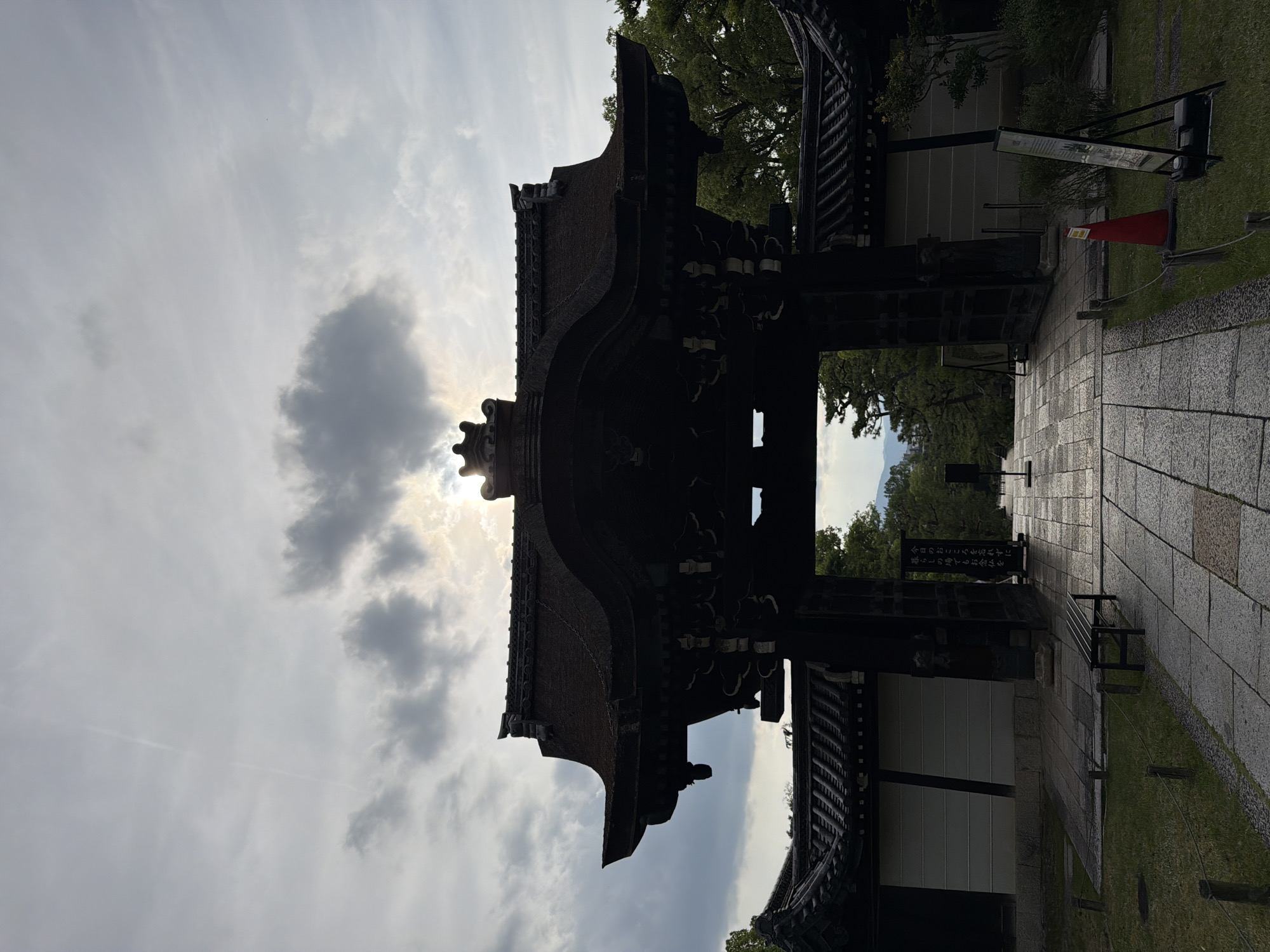 This image depicts the main entrance gate of Kodai-ji Temple in Kyoto, Japan. The grand, dark-wood gate is silhouetted against a bright, cloudy sky, with the sun partially obscured directly behind its peak. The gate features a prominent, multi-tiered, curved roof with traditional Japanese architectural details.
A stone-paved pathway leads directly through the center of the gate and continues into the distance. On the left side of the path, a dark metal bench rests on a patch of grass, bordered by a low rope fence supported by wooden posts. On the right side of the path, a black easel sign displays a white poster with Japanese text, stating: 「今日のこころをお忘れずに」 (Don't forget today's heart/spirit) and 「暮らしの場ではたらく人仏を」 (People who work in daily life). Further down the path on the right, a red traffic cone and another display stand are visible.
Beyond the gate, a view opens to a landscape of green trees and distant hills or mountains under a bright, overcast sky. A faint, thin contrail is discernible in the upper left portion of the sky. No individuals are present in the scene. The lighting emphasizes the silhouette of the gate and the path, creating a high-contrast image.
