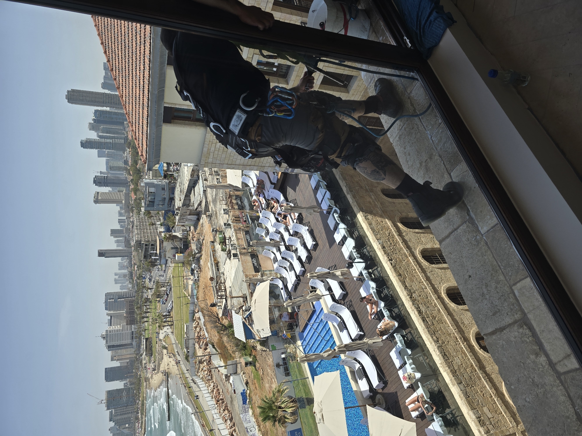 A window washer rappels down the side of a light-colored stone building, as seen from inside a window. The building appears to overlook a coastal scene. A swimming pool is visible, surrounded by numerous white lounge chairs with people sunbathing. Beyond the pool area, a beach with waves is seen. Further in the background is a dense urban skyline, suggesting a city.
Based on the style of buildings and layout, this location is likely in Tel Aviv, Israel.