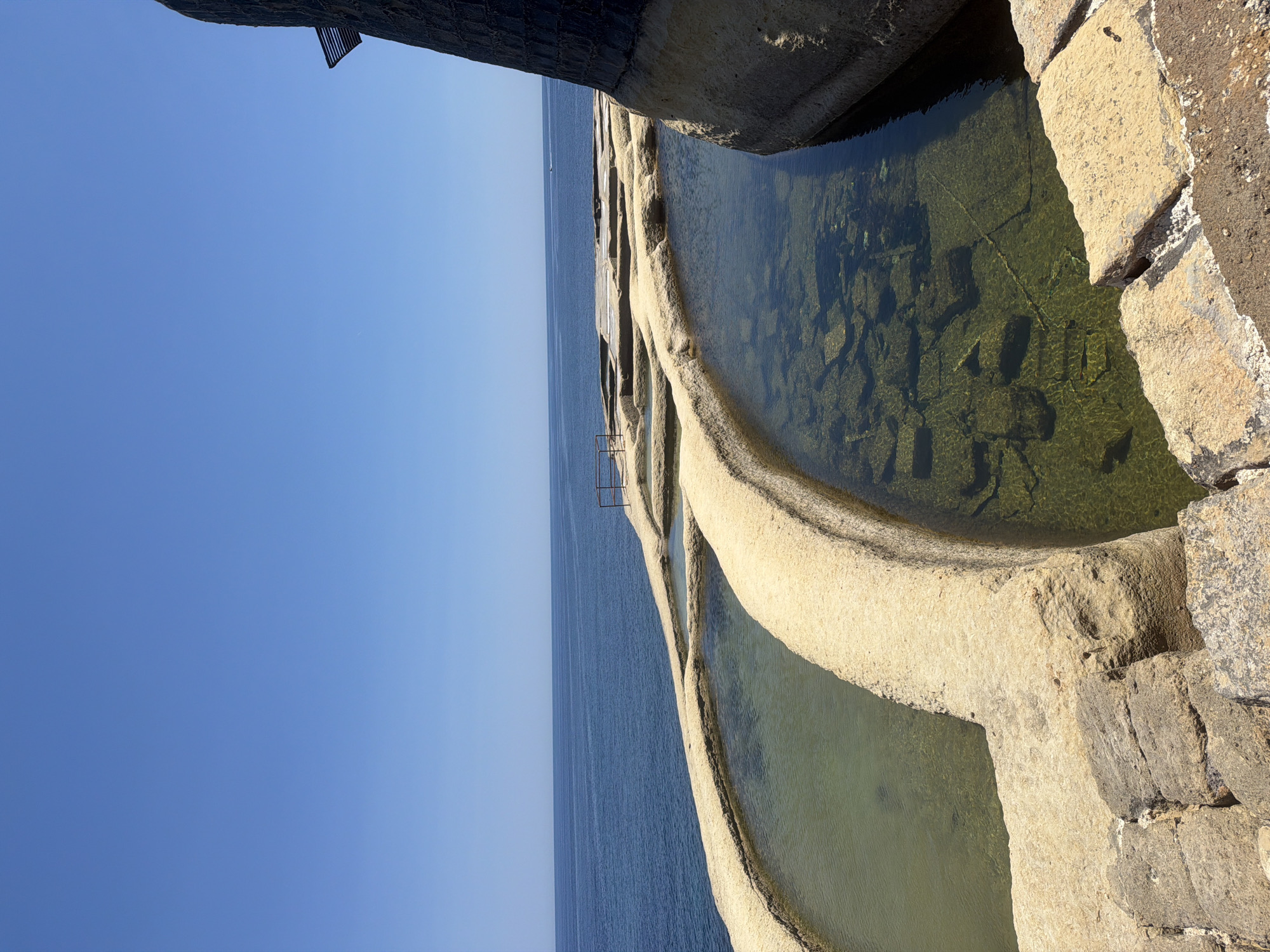 The image depicts a series of shallow, rectangular rock-cut pools situated along a coastline. These pools, likely salt pans, are filled with clear water, revealing the rocky and uneven bottom of each basin. The surrounding rock formations are weathered and appear to be limestone, typical of the Maltese landscape. In the foreground, there are rough-hewn stone steps leading down to one of the pools.
To the right of the pools, a dark, stone-built structure with a slatted opening rises vertically, casting a shadow on the adjacent rock. This structure could be part of a historical salt processing facility or a lighthouse. Beyond the pools, the deep blue expanse of the Mediterranean Sea stretches to the horizon under a clear, bright blue sky. A railing is visible on the edge of one of the pools, suggesting a designated pathway or viewing area. A small boat can be seen in the distance on the sea. The overall scene is one of a coastal natural and historical site, likely used for salt production.
The location is identified as the Salt Pans of Marsalforn, located on the northern coast of the island of Gozo, Malta. This area is known for its traditional method of salt harvesting, where seawater is channeled into these rock-cut pans and then evaporated by the sun and wind.