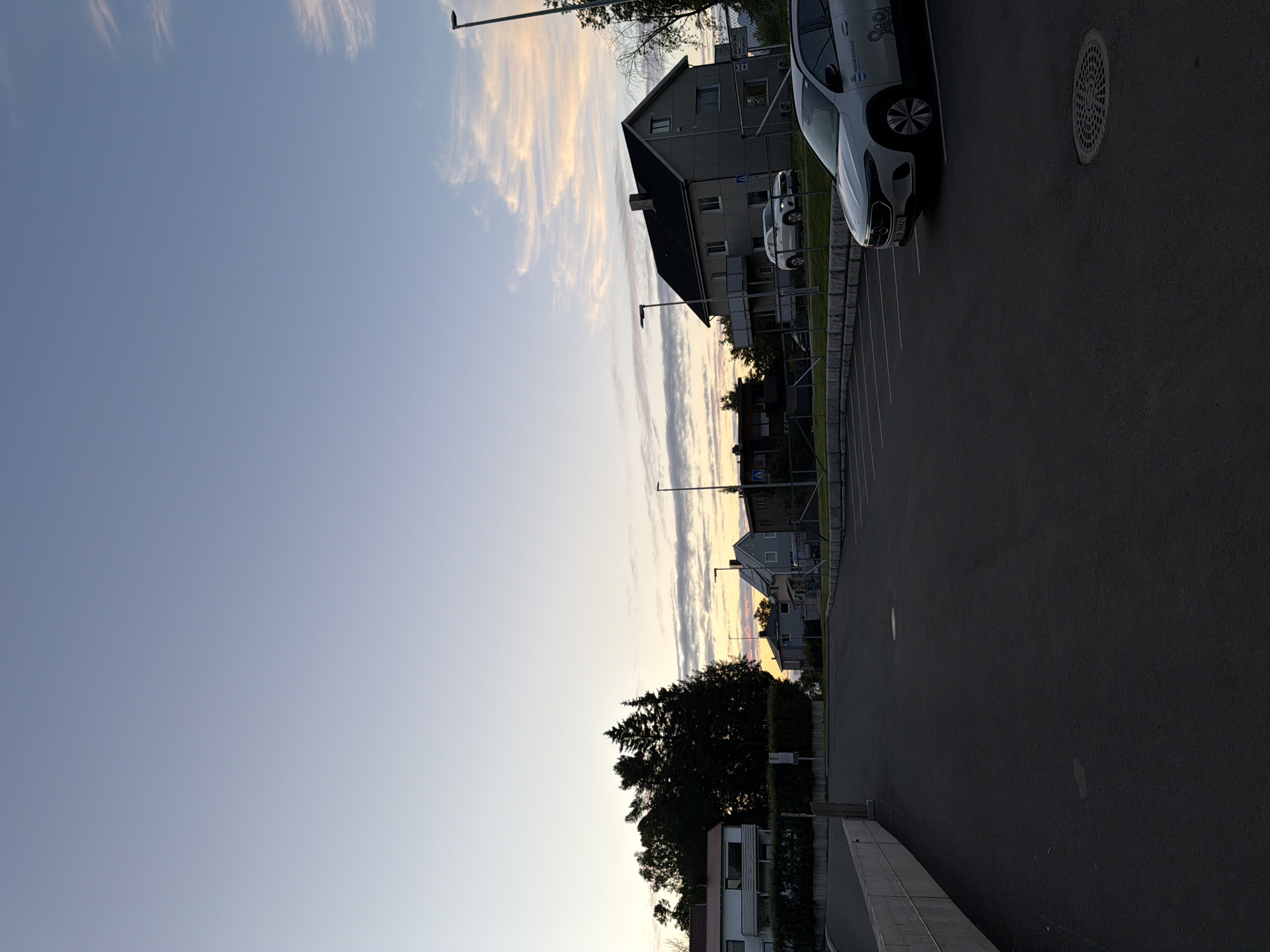 The image depicts an outdoor scene in the city of Harstad, Norway, during either dawn or dusk. The sky is a gradient of pale blue transitioning to pinkish hues near the horizon, with scattered wisps of clouds. In the foreground, a dark asphalt parking lot is visible, marked with white parking lines. A white electric vehicle with a license plate "FN 36471" is parked in the lot. Behind the parking lot, a low stone wall separates it from a grassy area. Several residential buildings, appearing to be houses, are visible in the middle ground, varying in architectural style and color. A dense cluster of dark green trees stands between some of the buildings. Streetlights are present, casting a faint glow. The overall impression is of a quiet, suburban area at the beginning or end of the day.