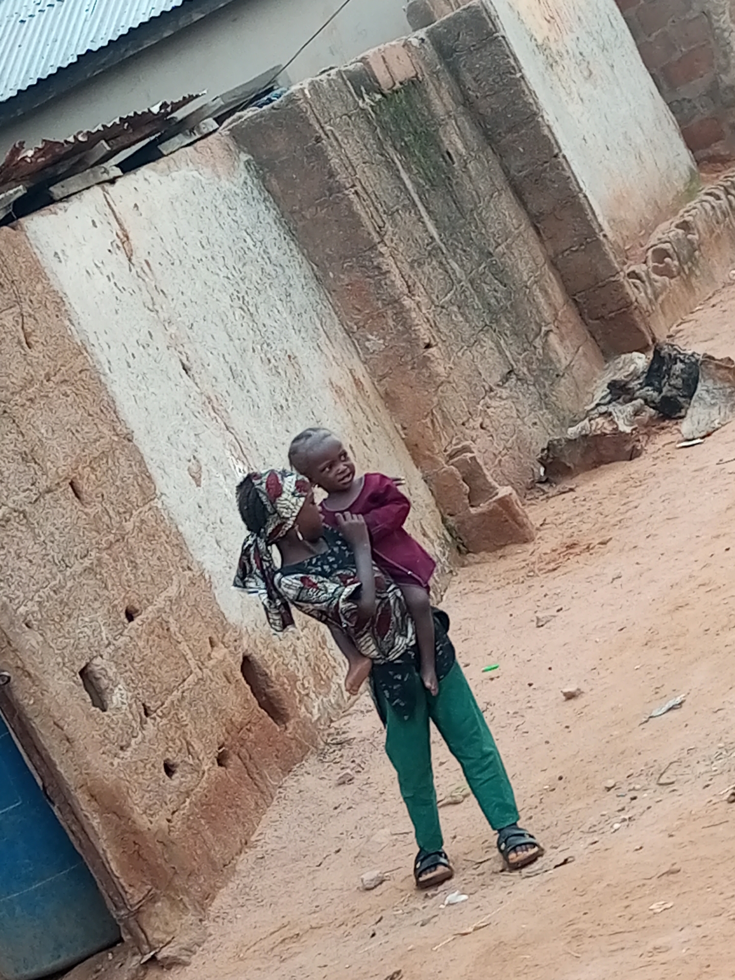 In Tumfure, Nigeria, a person carries a child. The person is wearing green pants and brown sandals. The child is wearing a maroon shirt and a patterned fabric around its body. They are standing on a dirt path next to a building constructed of rough, textured bricks. The building has a corrugated metal roof. The overall environment appears to be a rural or semi-urban setting.