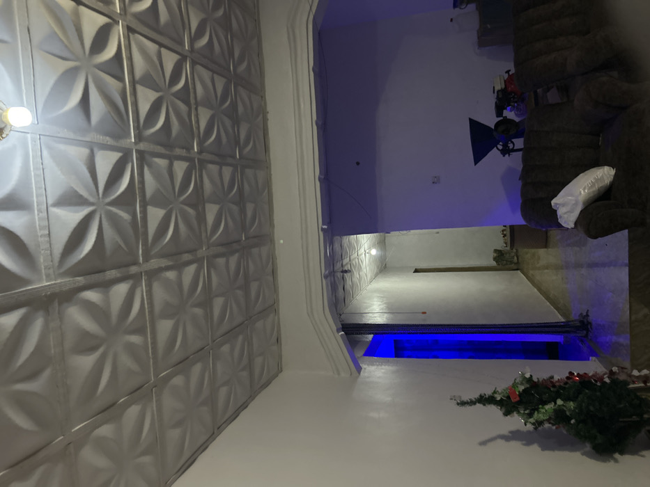 An indoor scene displays a gray decorative ceiling composed of geometric, floral-like patterns. The room is divided by a large archway. Beyond the archway is a living room area with a brown couch and a white pillow. To the right, a partial Christmas tree lies on a white floor, bathed in blue light. The lighting creates a cool, somewhat sterile ambiance, contrasting with the warmth of the living room section.