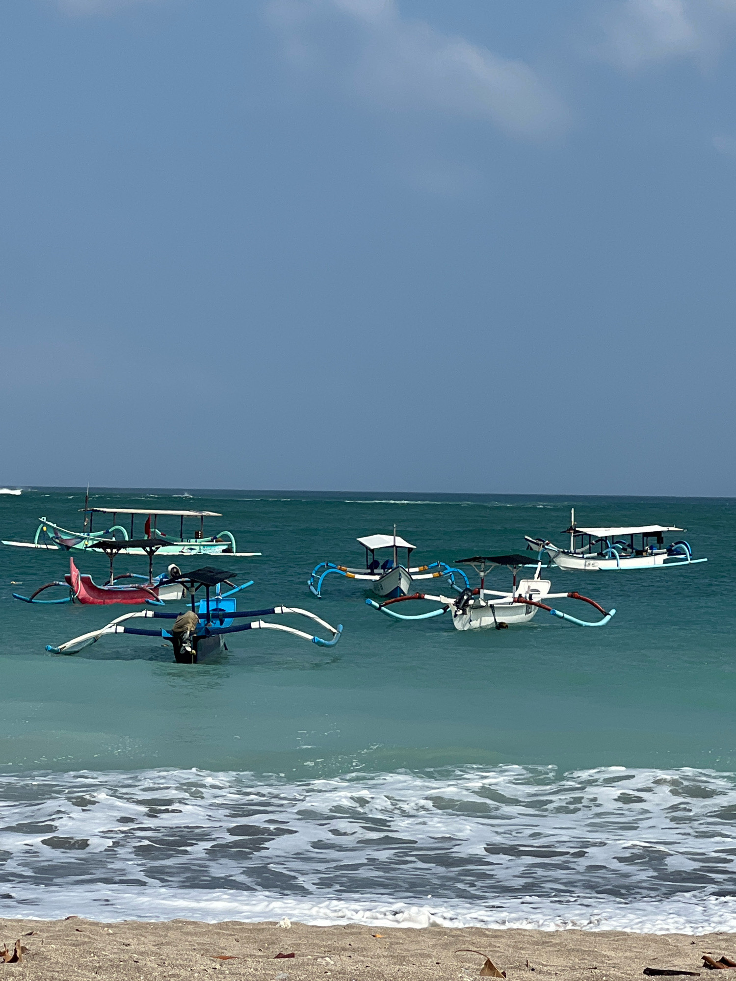 The image depicts a serene beach scene in Kuta, Indonesia, with several traditional Balinese outrigger boats (jukung) floating in the clear turquoise water. The boats are of various sizes and colors, including white, red, and green, with distinctive bamboo outriggers extending from their sides. Some boats are equipped with canopies, offering shade from the sun. The sea is calm, with gentle waves lapping at the shore, and the sky is a pale blue with scattered white clouds. The beach itself is made of light-colored sand, with some dry leaves and debris scattered on it. There is no visible human activity apart from a single person sitting in one of the boats. The overall atmosphere is peaceful and tranquil, characteristic of a tropical beach destination.