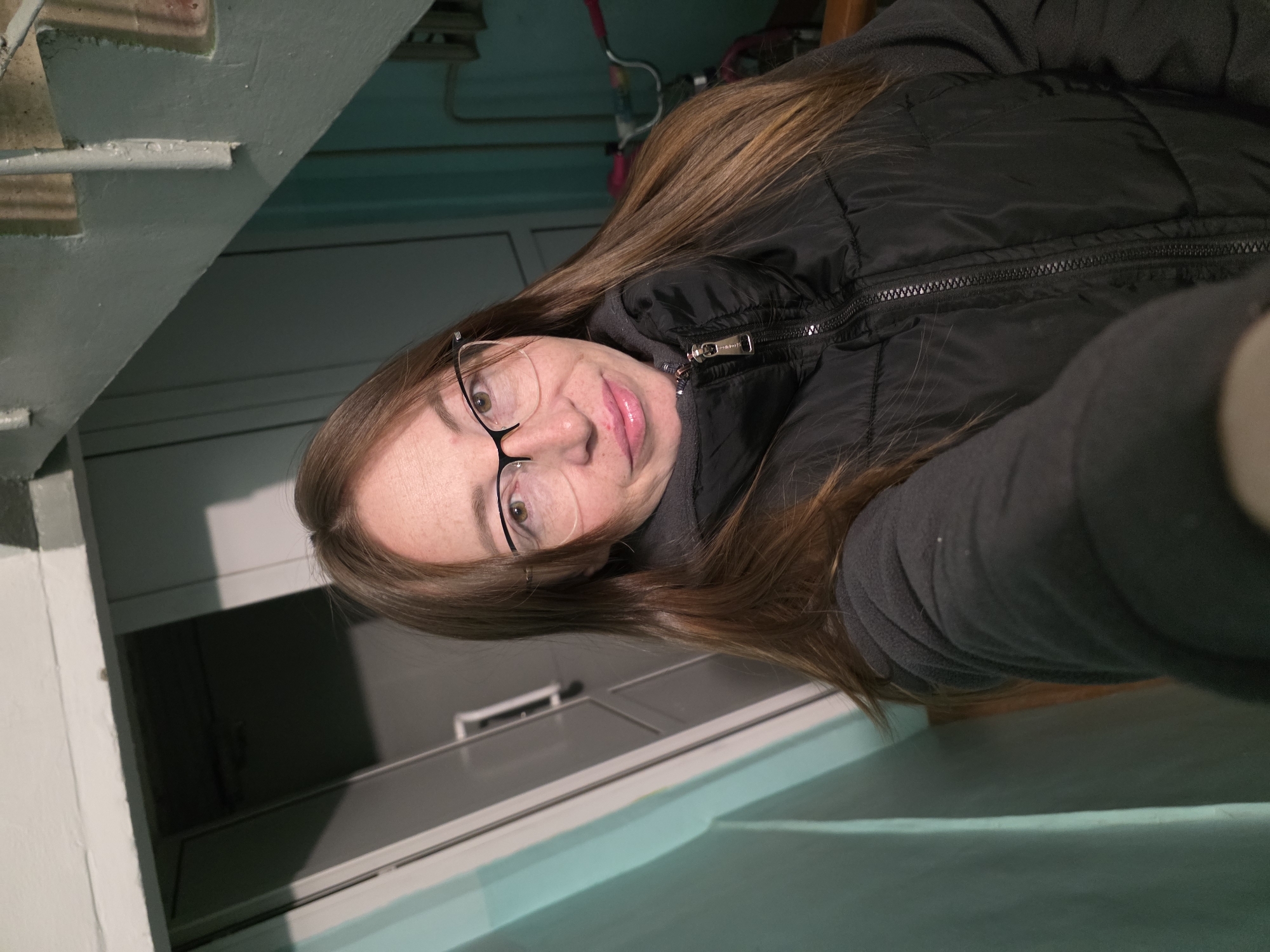 A woman with long, reddish-brown hair and clear-framed glasses takes a selfie, causing the image to appear rotated counter-clockwise. She has light skin with freckles and is smiling softly, looking directly at the camera. She wears a black quilted puffer vest over a grey long-sleeved top.
The setting is an indoor space in Chaikovsky, Russia. To the woman's left, a light green or turquoise painted wall or structural element, possibly part of a staircase, is visible. Behind her, there are light-colored cabinets or shelves, one appearing open with a dark interior. Above her, more of the light green structure is present, along with hints of thin red and white elements, possibly pipes or wiring in the background. The lighting is artificial and appears somewhat dim. A small, unreadable logo is visible on the zipper pull of her vest. The scene captures an informal, candid moment.