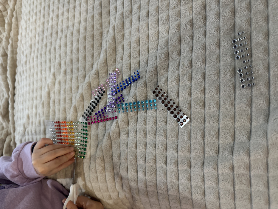 The image displays a close-up, high-angle view of a crafting activity taking place on a light beige or light brown textured fabric surface, likely a blanket or bedspread. In the upper left quadrant, the left forearm and hands of an individual, wearing a light purple long-sleeved top, are visible. The left hand holds a clear plastic strip of small, round, self-adhesive rhinestones arranged in vertical lines of red, orange, and green. The right hand manipulates a pair of small, white-handled scissors, positioned to cut this rhinestone strip.
Various other clear plastic strips of self-adhesive rhinestones are scattered across the fabric surface. These include black, pink, purple, blue, cyan, and dark brown rhinestones, with some strips appearing partially cut or individual rhinestones detached. A small collection of silver or clear rhinestones is loosely arranged in the lower right portion of the frame. The scene indicates an indoor domestic environment where a creative project is underway. The precise location within the city of Varna, Bulgaria, cannot be determined from the visual evidence in the image.