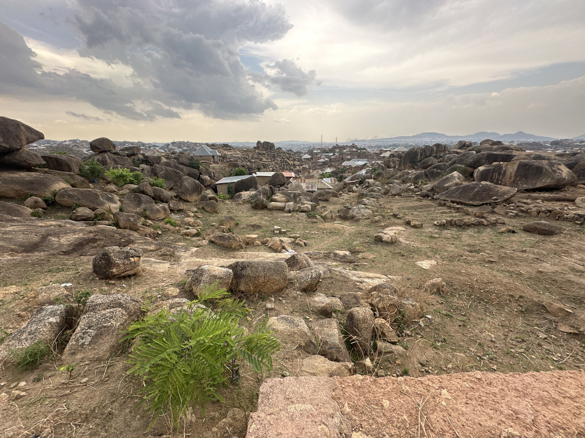 The image depicts a rocky landscape with scattered dwellings in Jos, Nigeria. The foreground features a mix of exposed earth, loose rocks, and sparse vegetation. A small, green bush is visible in the lower left corner. Larger, rounded boulders and rock formations dominate the middle ground. Houses with corrugated metal roofs are interspersed among the rocks, suggesting a settlement integrated within the natural landscape. In the distance, a cityscape is visible, with buildings extending towards a horizon marked by rolling hills and a cloudy sky. The overcast conditions cast a muted light over the scene.