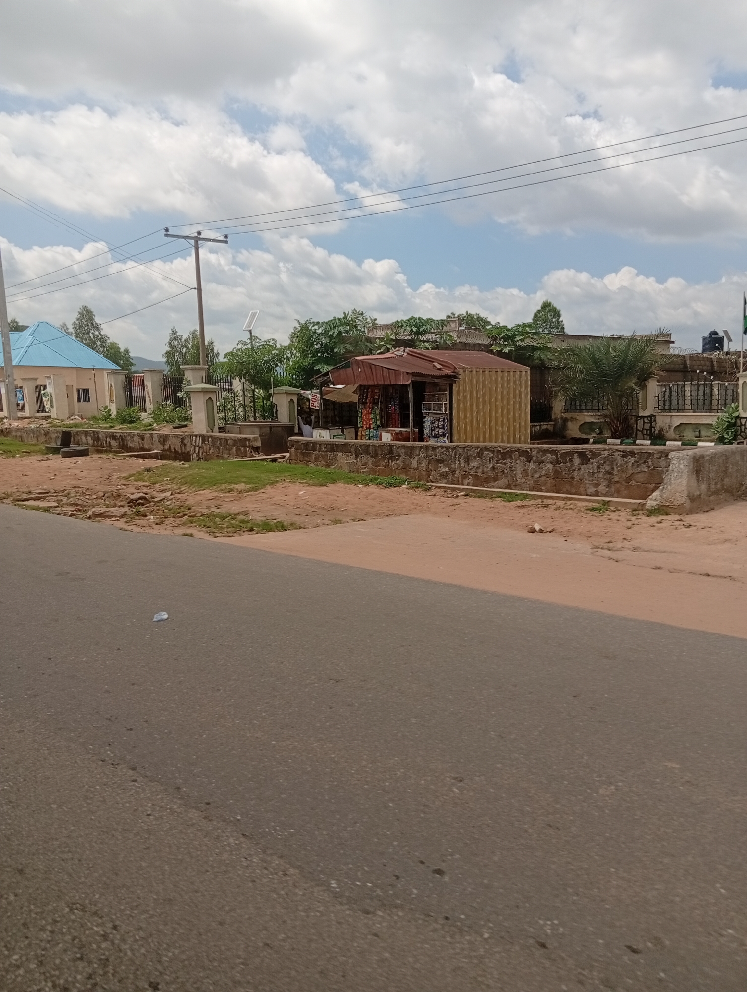 The image shows a street scene in Jalingo, Nigeria, on a cloudy day. In the foreground, a paved road is visible, with a plastic bag lying on the surface. To the left, a light blue building with a light blue roof stands behind a low, rough-stone wall. Further to the left, a utility pole with wires runs across the frame. In the middle ground, there is a small market stall with a corrugated metal roof, displaying various goods. Adjacent to the stall is a corrugated metal container. A wall made of rough stones and concrete runs along the front of the market area. Behind the wall, there are various trees and foliage. In the background, there are more buildings and the sky is filled with white, fluffy clouds. A water tank is visible on top of one of the buildings. A Nigerian flag can be seen on a pole at the far right of the image. There are no people clearly visible in the scene.