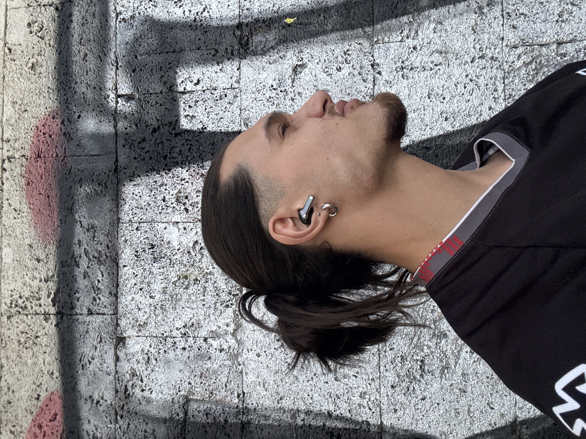 A man is lying on a concrete surface that is partially painted with graffiti. The man has dark hair pulled back into a ponytail, facial hair, and earrings and a Bluetooth device in his left ear. He is wearing a black shirt. The concrete surface appears to be a wall. The location is the city of Sofia, Bulgaria.