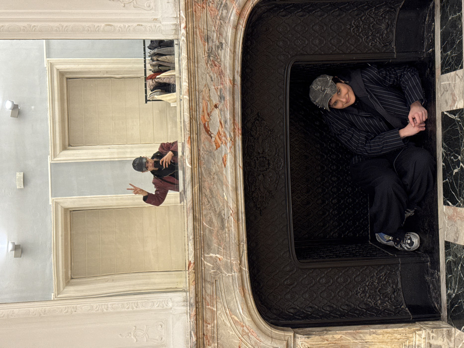 An interior scene in Sofia, Bulgaria, features two individuals in a grand room with ornate architectural details. In the foreground, a person is lying on their left side within a dark, unlit fireplace opening, which is adorned with patterned cast iron. This individual wears a dark pinstripe jacket over a dark top, dark wide-leg pants, and a distressed camouflage-patterned baseball cap. Their left foot wears a multi-colored sneaker, and their hands are clasped. They are looking directly at the viewer with a slight smile.
Above the fireplace, a large mirror is mounted, featuring a light-colored, ornate frame and reflecting the room behind the viewer. The fireplace mantel is constructed from polished marble with white, grey, and reddish-brown veins.
Reflected in the mirror, a second person, presumably the photographer, is visible. This individual is wearing a dark cap, a dark red jacket, and a dark scarf, holding a smartphone with their left hand to take the picture. Their right hand is raised, displaying a peace sign. Behind the reflected person, a light blue wall is visible, featuring two white, ornate doorframes with closed light-colored panels. A clothing rack with several garments hangs to the right of the reflected person. Two square-based wall lights are visible on the upper section of the reflected wall. The floor in front of the fireplace consists of polished black and white marble tiles arranged in a checkerboard pattern.