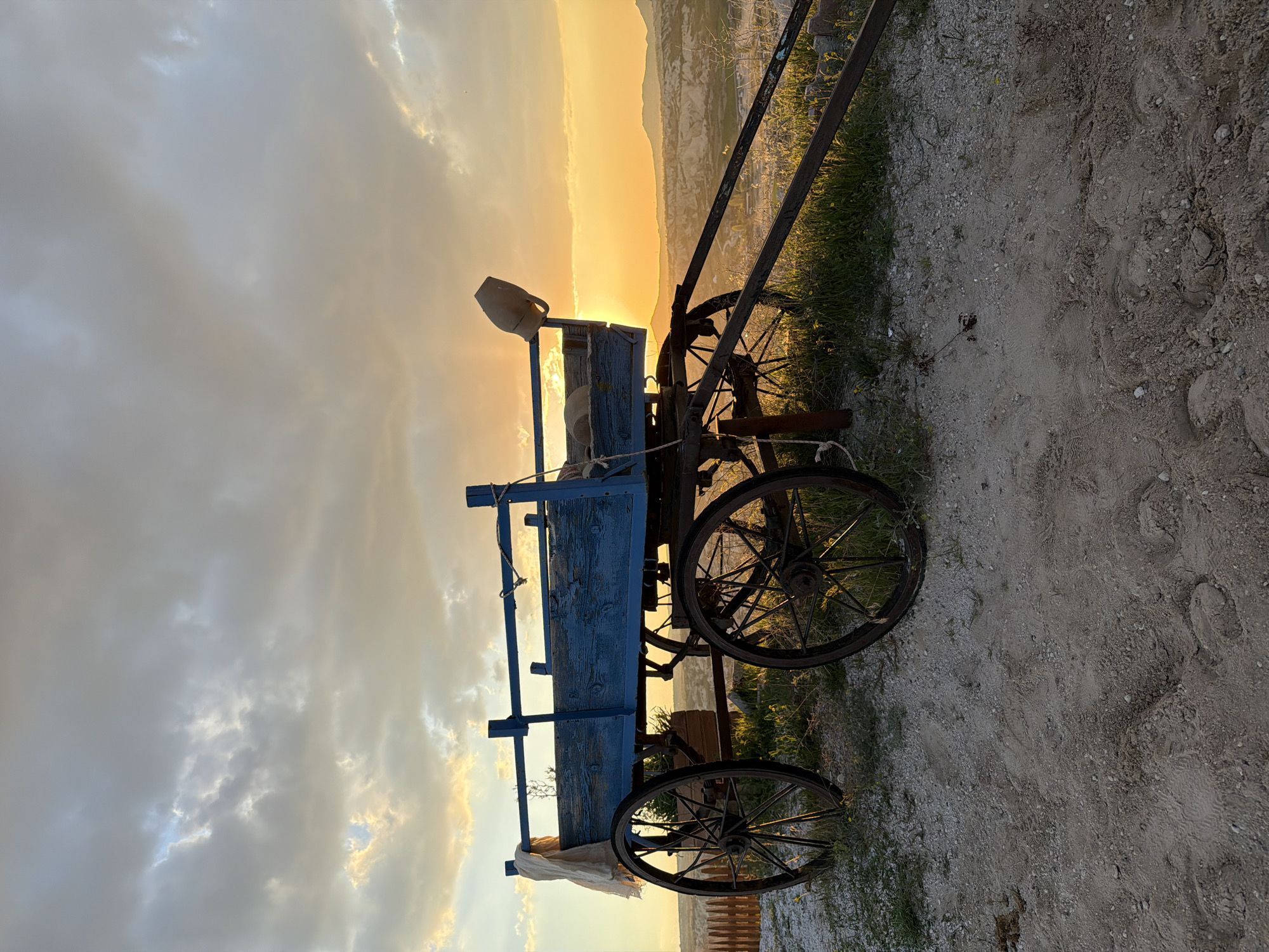 The image displays an old, weathered wooden cart, painted in a faded blue, set against a vibrant sunset. The cart features large, spoked metal wheels. A light-colored jug or pitcher rests on its side, and a piece of white fabric is draped over another part of the frame. The ground in the foreground is a dirt path with scattered small rocks, sparse green grass, and small yellow wildflowers. In the midground and background, undulating hills or distinctive rock formations are visible, consistent with the Göreme region in Turkey, softly silhouetted by the intense, golden light. The sky transitions from a brilliant orange and yellow at the horizon, where the sun is setting, to softer grey and white clouds higher up. The time of day is clearly sunset, characterized by the warm, directional light and vivid colors. No people are visible, and there is no discernible text within the frame. The scene conveys a sense of quiet rural beauty at dusk.