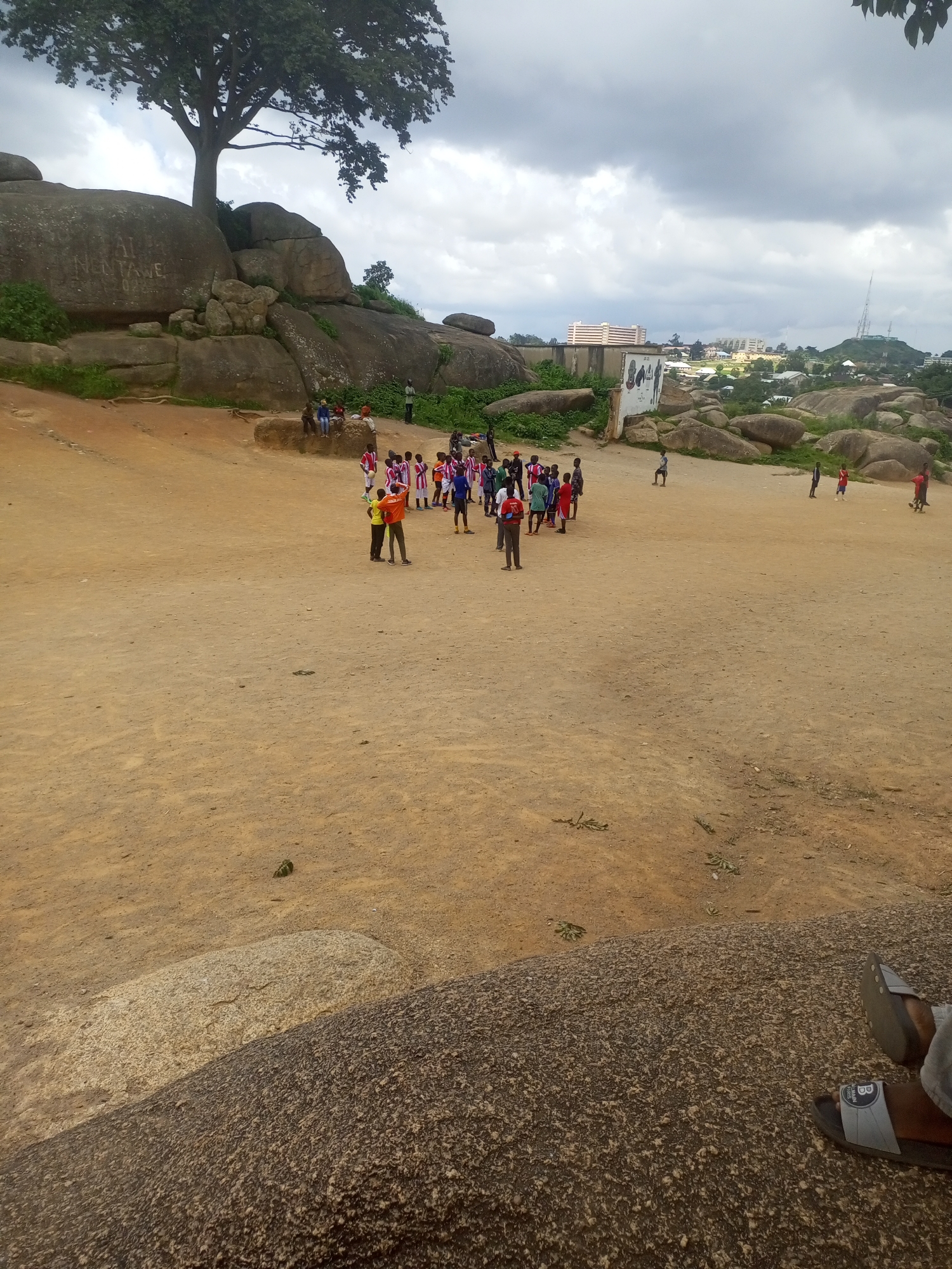 The image depicts a gathering of children, seemingly engaged in a sporting activity, on a dusty, open field surrounded by large rock formations. A prominent tree with a full canopy stands behind the rocks to the left. In the background, the cityscape of Jos, Nigeria, is visible, with buildings and communication towers dotting the horizon under a cloudy sky. Some of the children are wearing matching red and white attire, suggesting a team or organized event. The terrain is uneven, with scattered rocks and sparse vegetation. The overall scene conveys a sense of community engagement in an outdoor recreational setting within the unique rocky landscape of Jos.